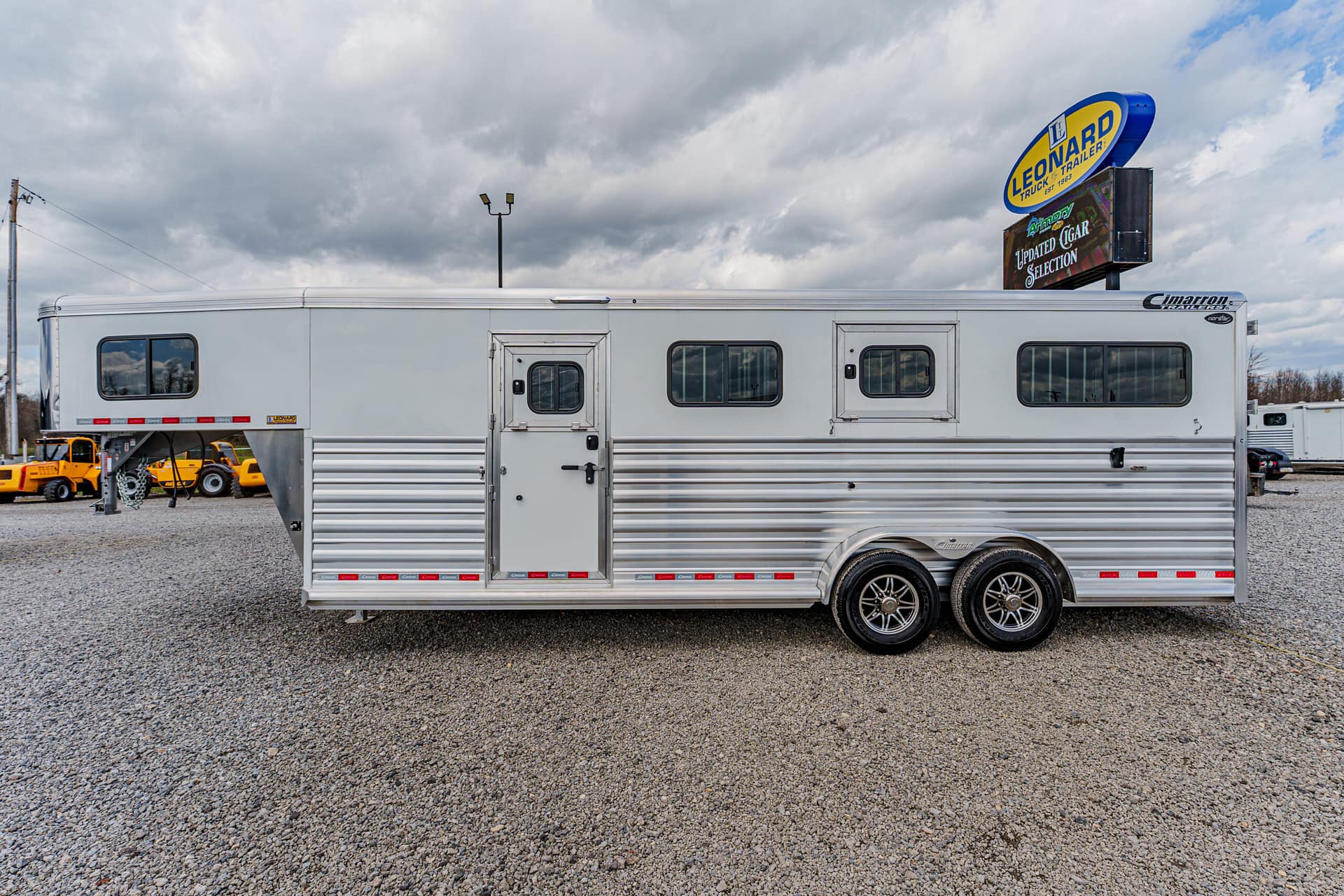 Silver aluminum horse trailer with two axles and multiple windows, parked on a gravel lot under a cloudy sky.