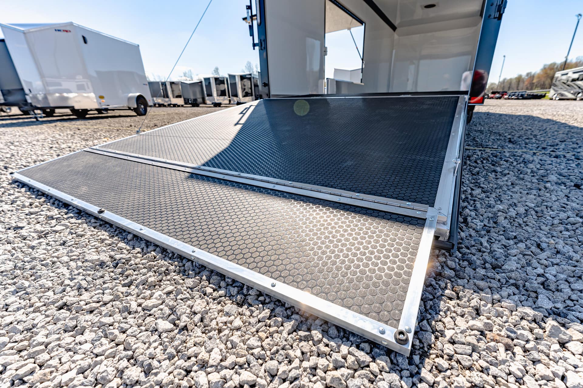 Close-up of a metal trailer ramp on a gravel lot, with other white trailers in the background.