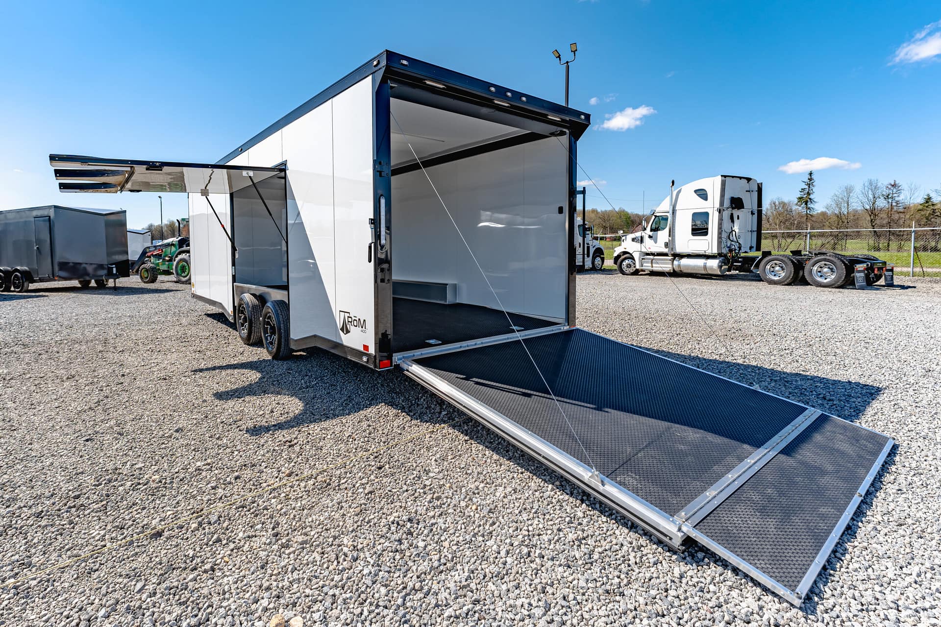 White utility trailer with a fold-out loading ramp on a gravel lot, with a semi-truck in the background on a clear day.