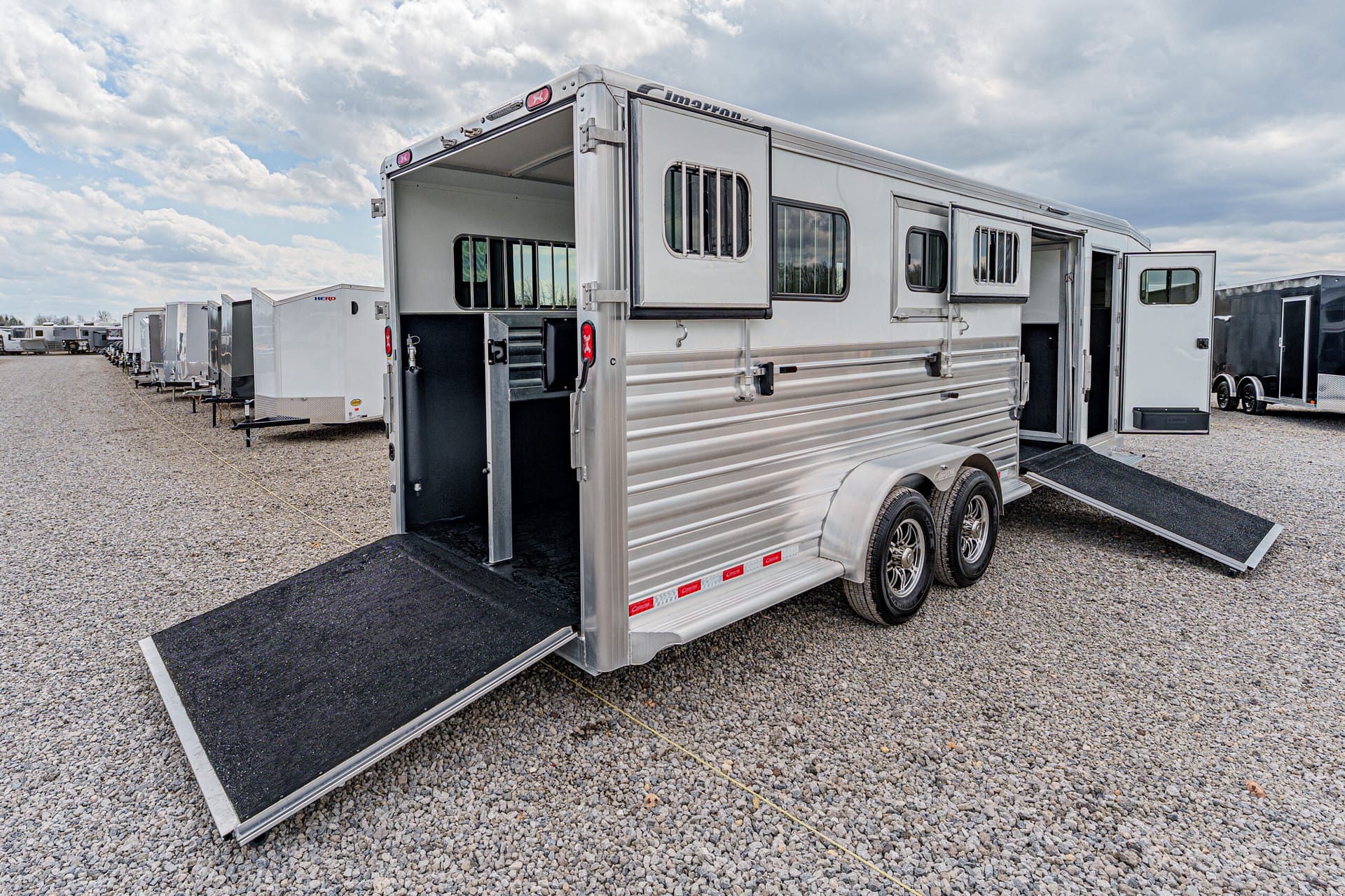 Aluminum horse trailer with two wide loading ramps lowered on a gravel lot, other trailers in the background