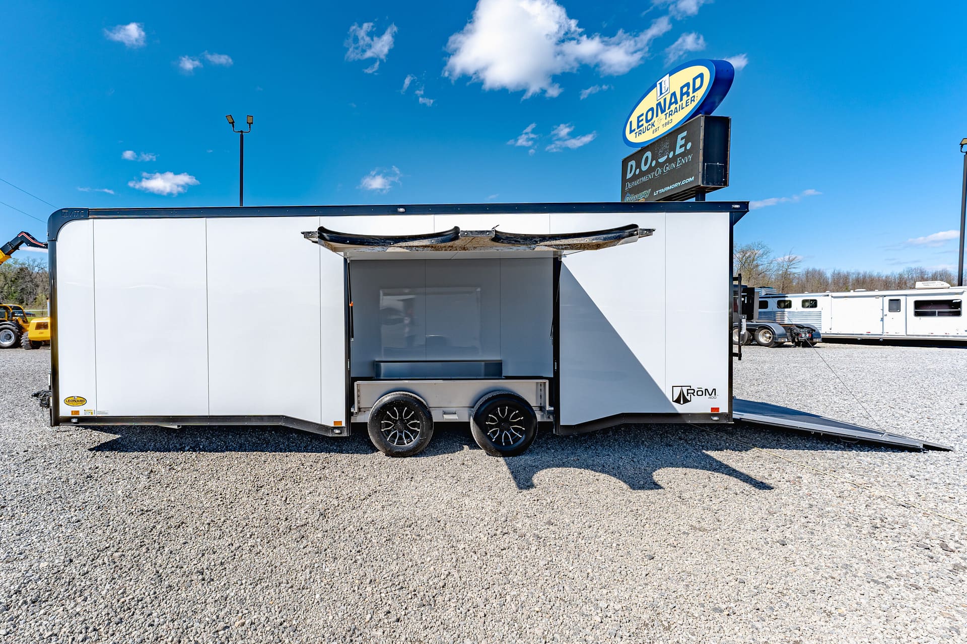 White enclosed trailer with its rear ramp lowered, parked on a gravel lot under a clear blue sky.