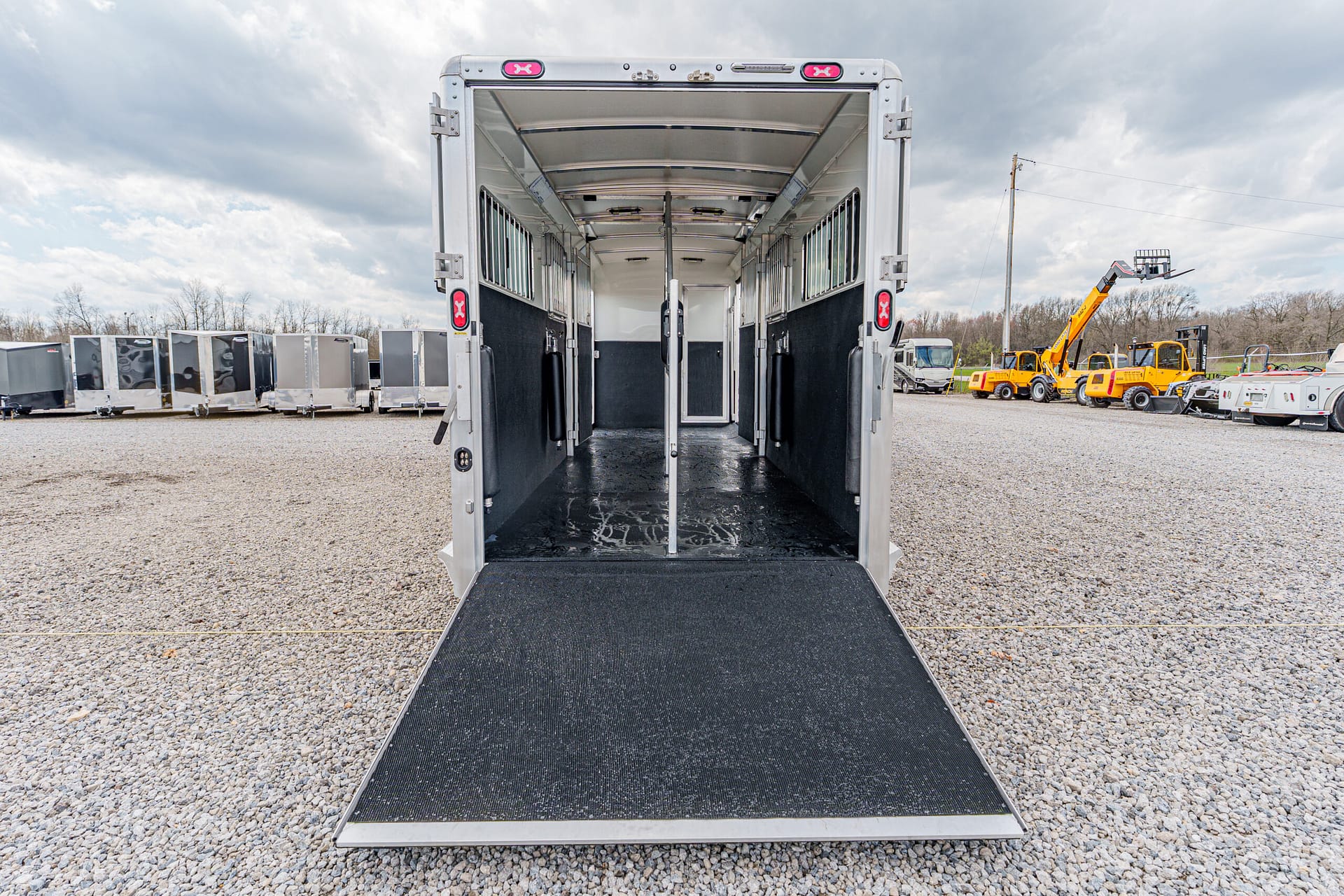Wide view of a silver horse trailer with ramp down, empty and ready for loading in a gravel lot.