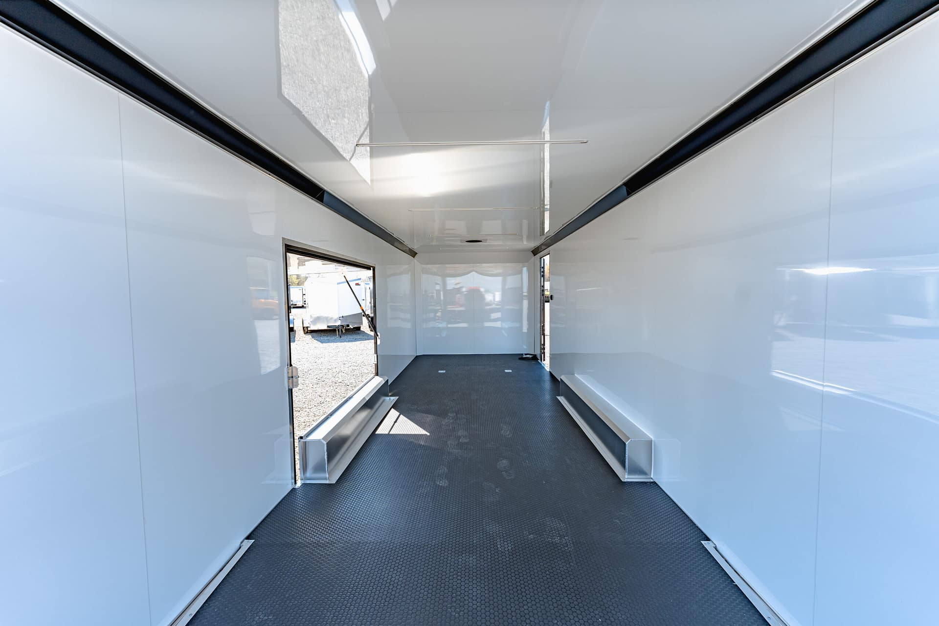 Interior of an empty white trade trailer with a black textured floor and open doors at both ends, outdoors visible through the fronts.