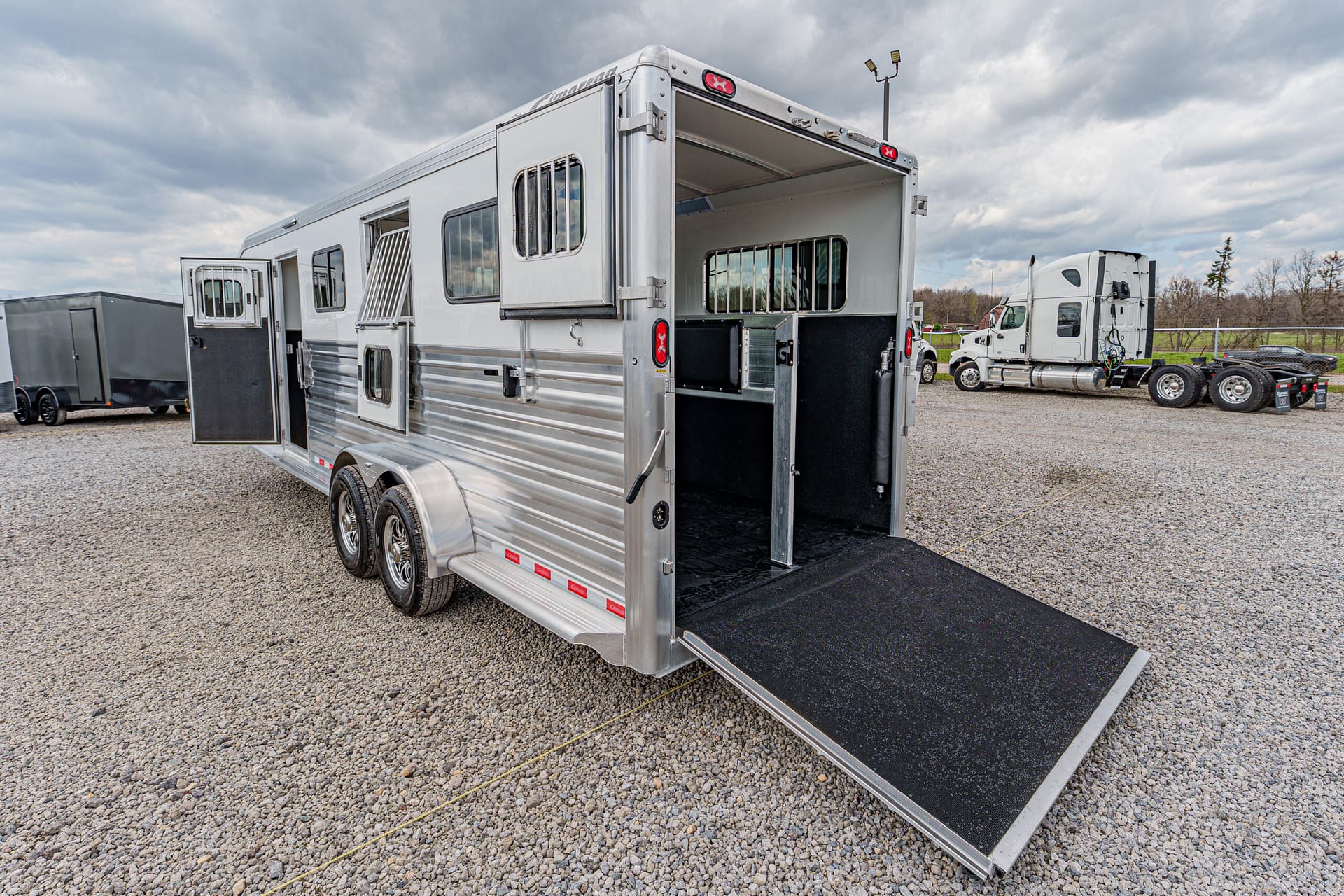 Silver aluminum horse trailer with its rear ramp lowered onto a gravel lot, showing interior stalls and barred window at the back.