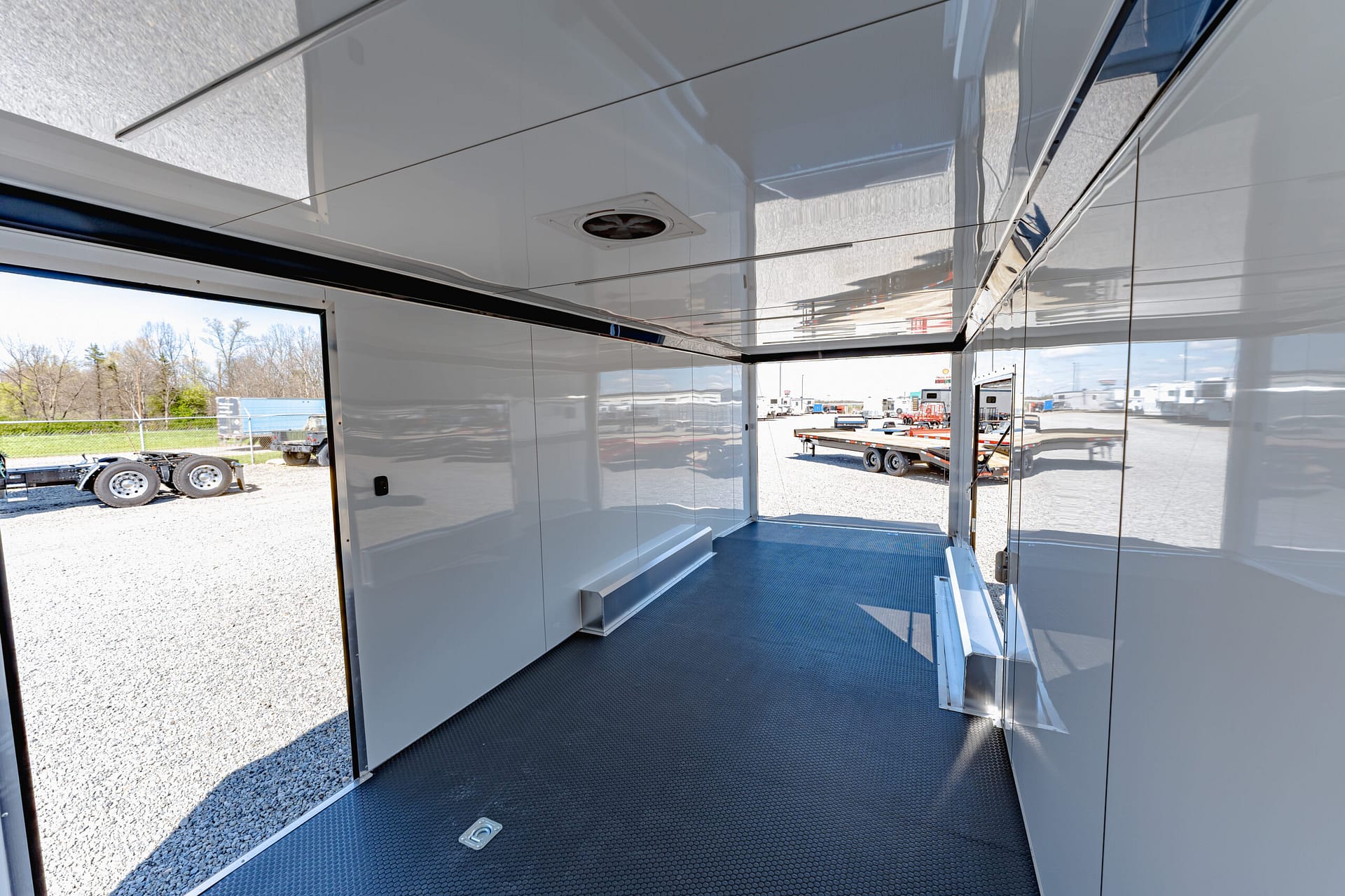 Reflection-filled aluminum trailer interior with a diamond-plate floor and an open side door showing a truck yard outside