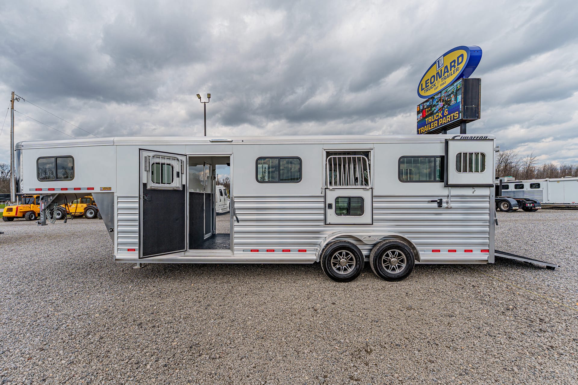 Silver horse trailer with open entry doors, parked on a gravel lot with a Leonard truck sign in the background (informational image).