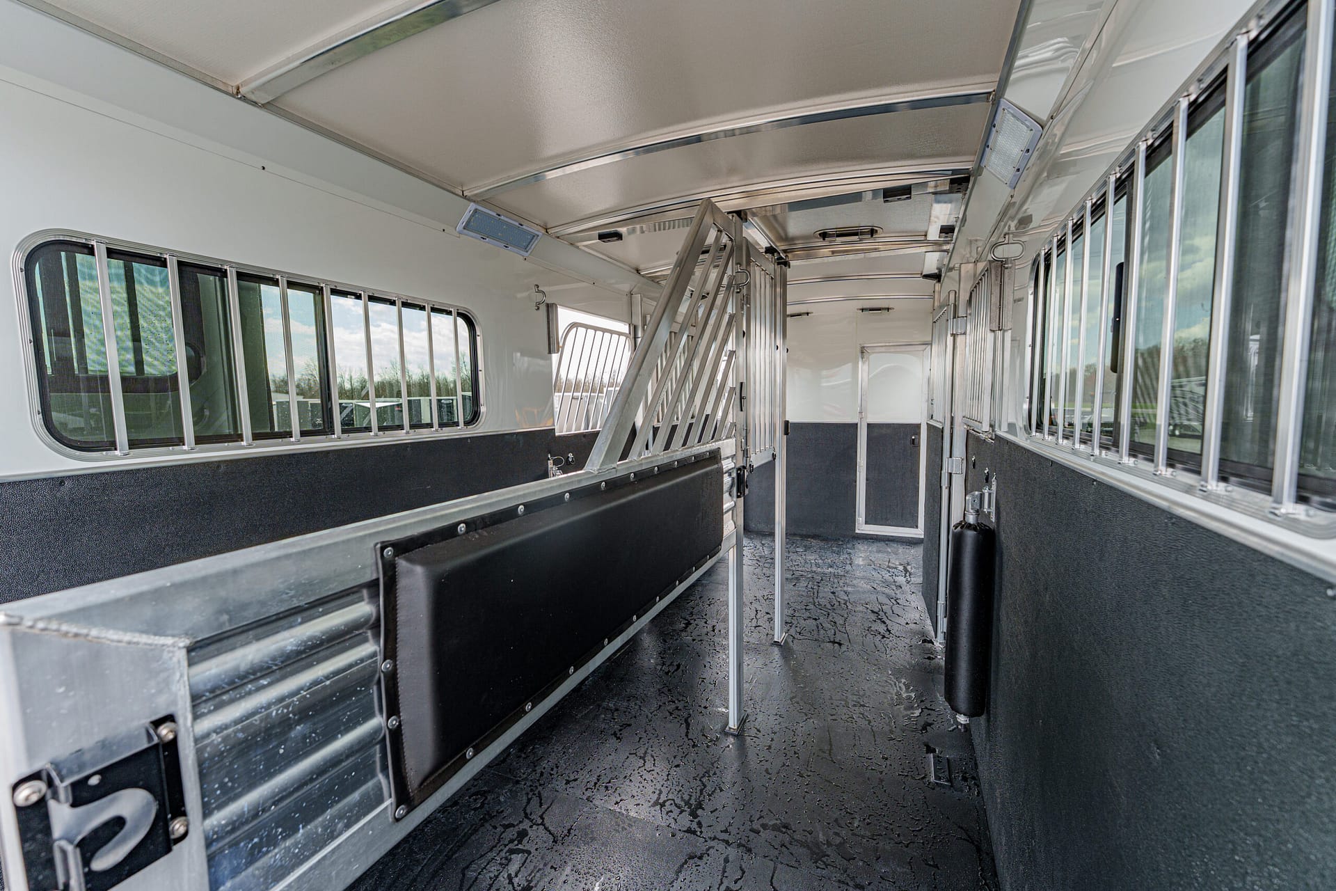 Interior of a horse trailer with partitioned stalls, padded divider along the left, metal bars on windows, and a textured rubber floor.