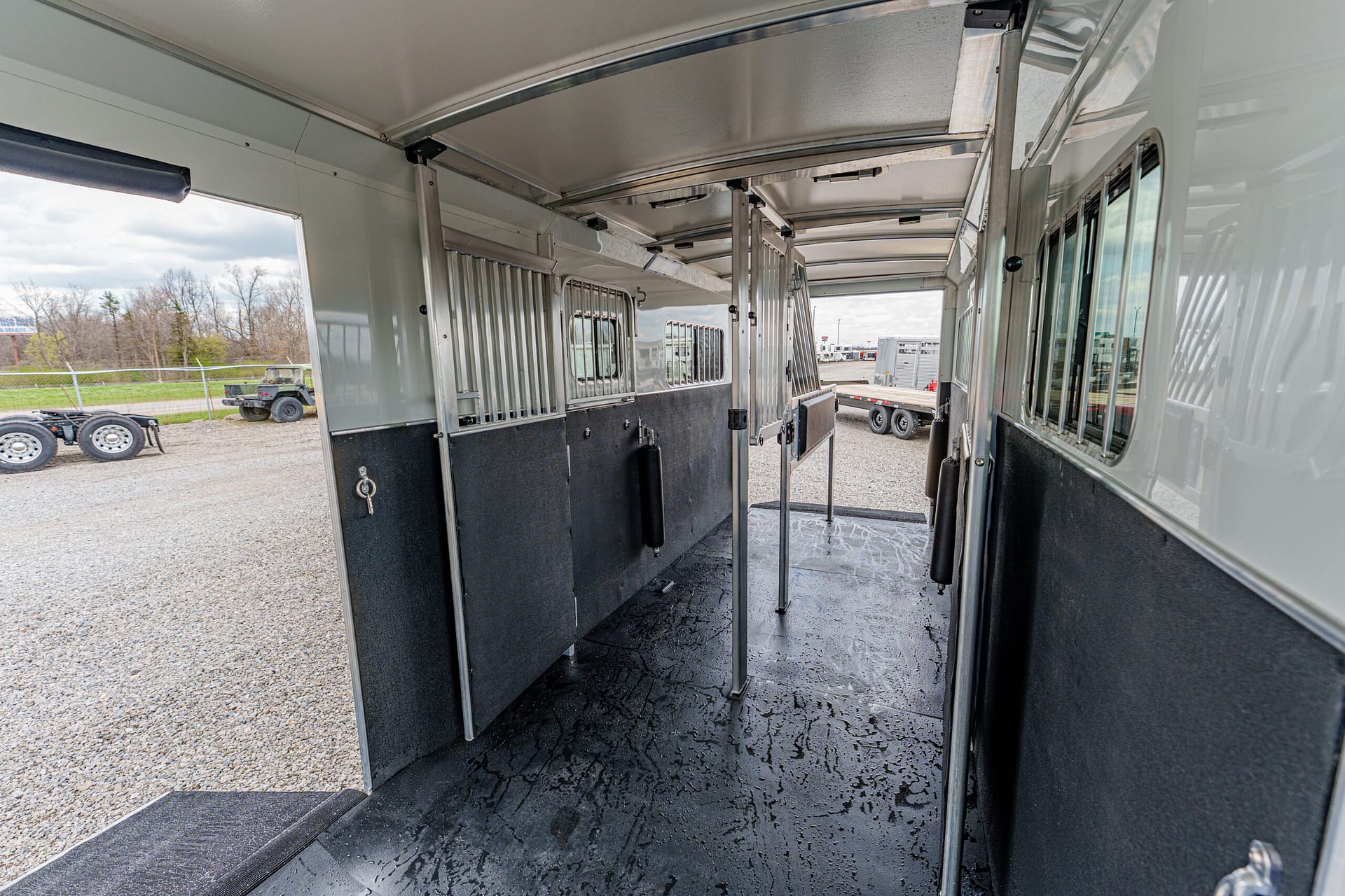 Interior of a horse trailer with metal divider stalls, black rubber flooring, and vertical partition bars; outside gravel lot visible through open side and rear doors.