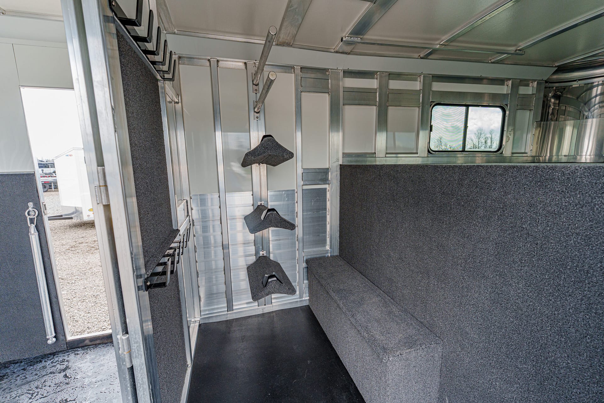 Interior of a metal horse trailer with padded gray walls, a bench, and hanging saddle racks; window shows trees outside.