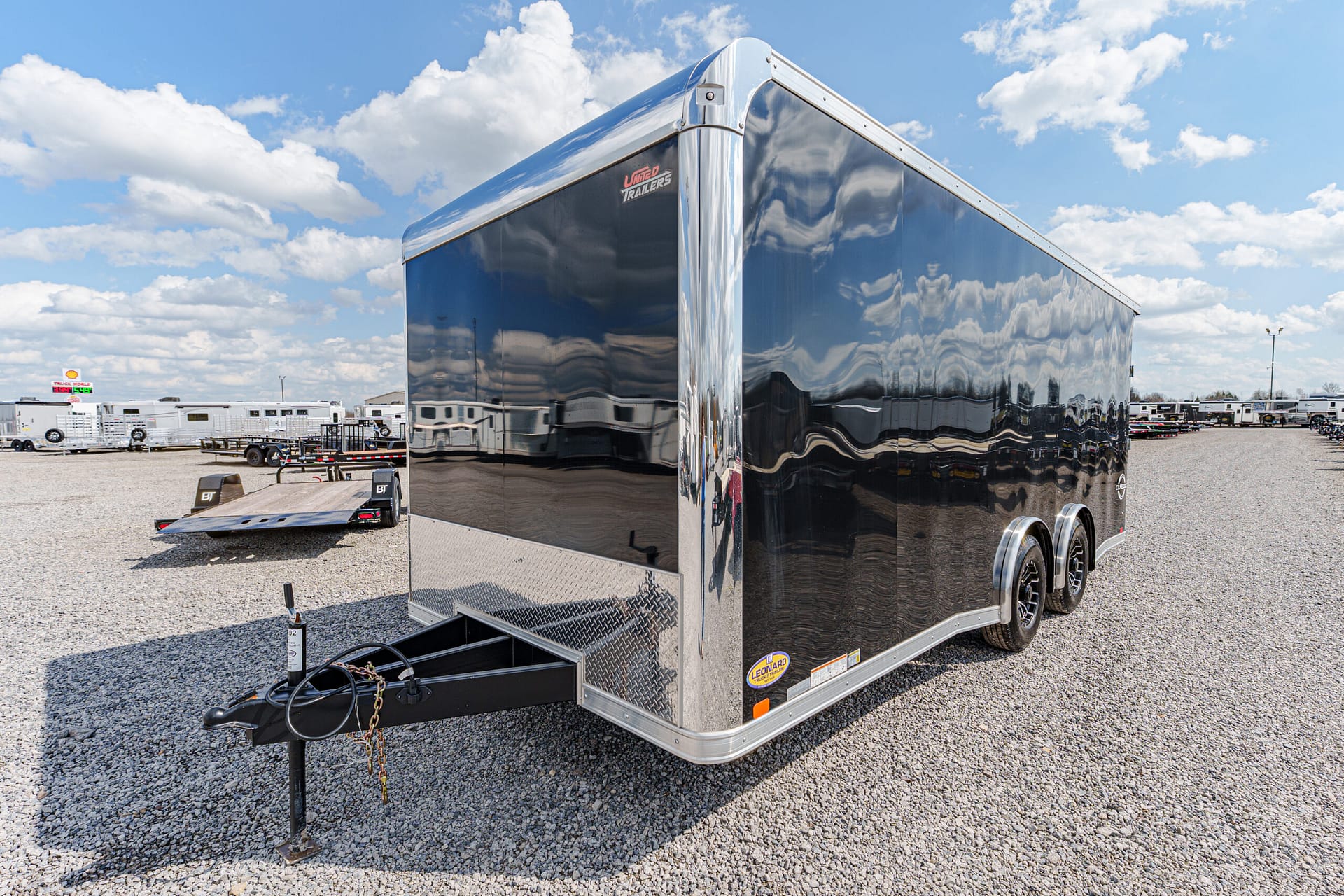 Glossy black enclosed cargo trailer with diamond-plate front and twin axles in a gravel lot, reflecting the sky and surrounding trailers.