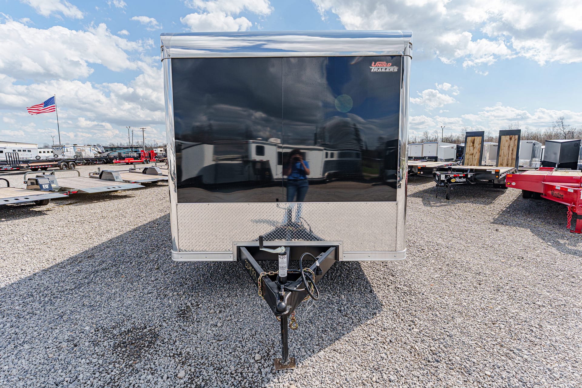 Front view of a shiny enclosed trailer on a gravel lot, hitch in foreground, reflection of photographer on the glossy door, with other trailers and an American flag in the background