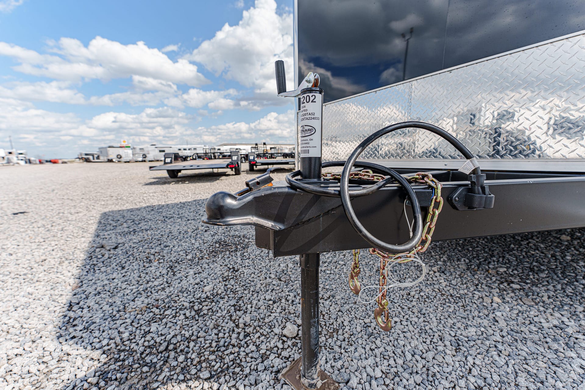 Close-up of a trailer hitch with safety chains and jack on a gravel lot, with other trailers in the background under a blue sky.