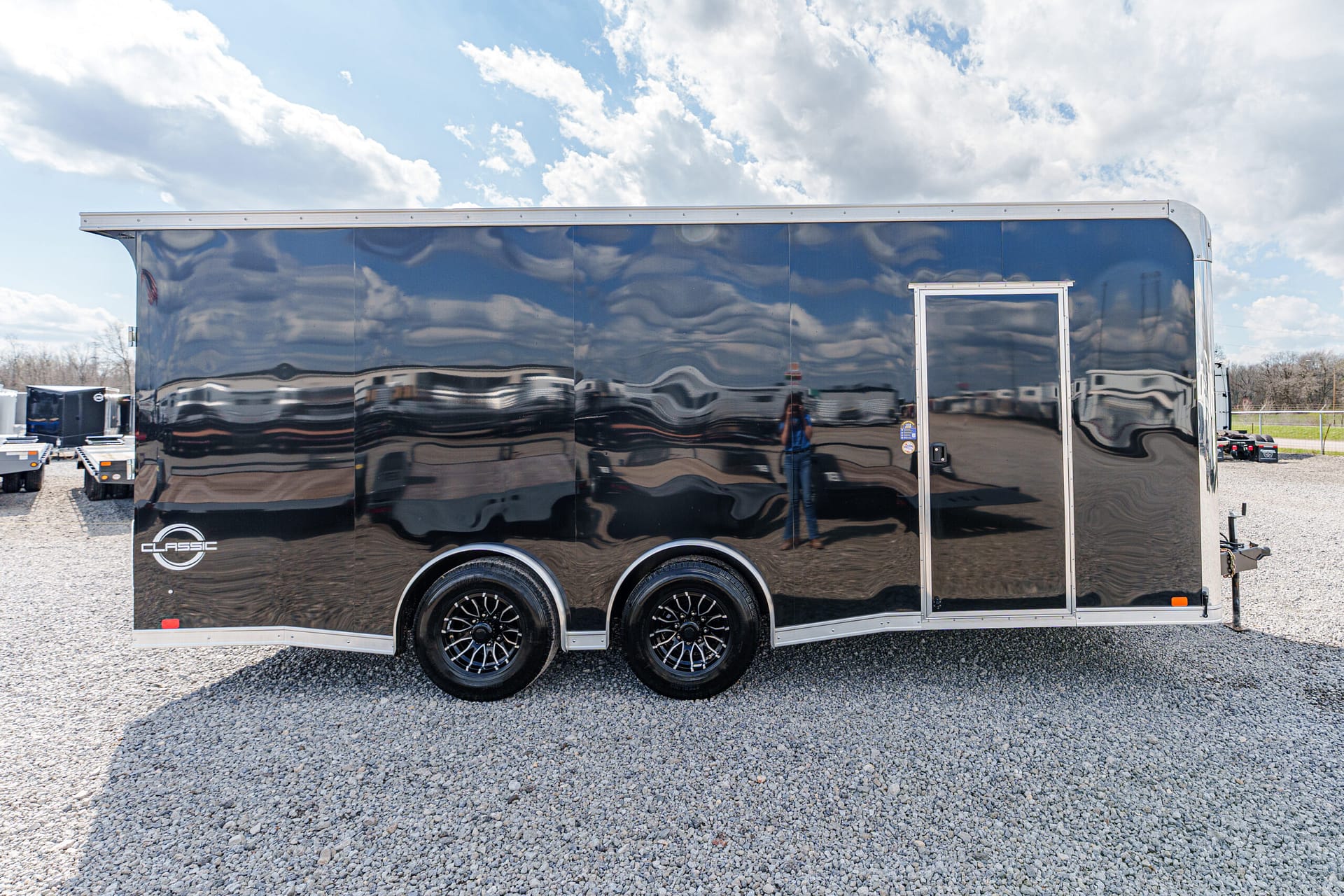 Shiny black enclosed trailer with two axles and a side door, parked on a gravel lot.