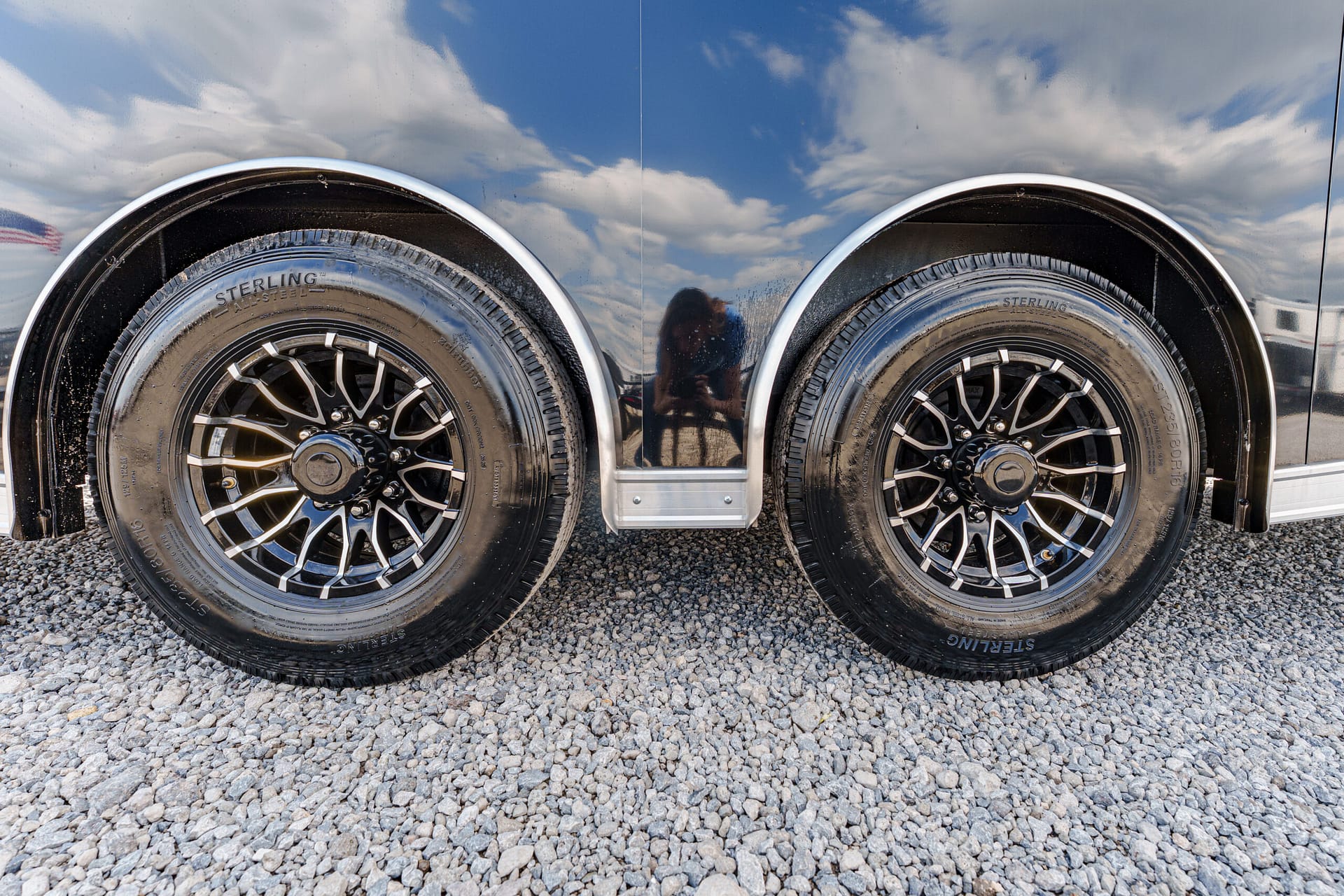 Dual black tires with chrome rims on a trailer; reflection of a person taking a photo on the shiny body.