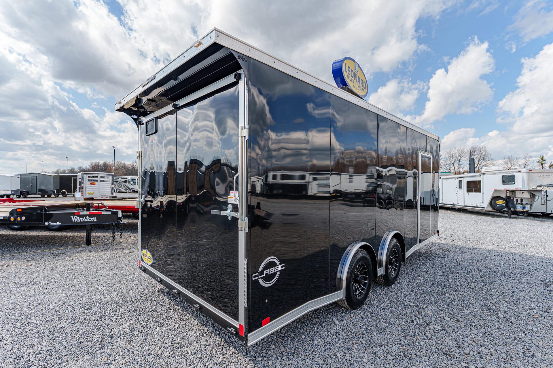 Black enclosed dual-axle cargo trailer with chrome trim and Leonard sign on top, parked in a dealership lot.