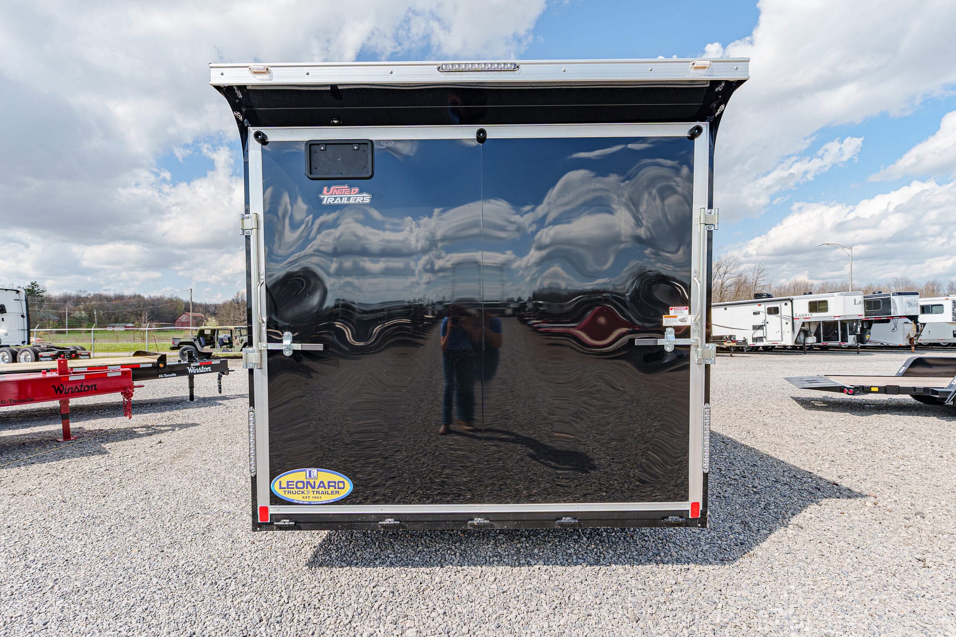 Rear of a glossy black enclosed trailer with silver trim and Leonard trailer sticker on a gravel lot.