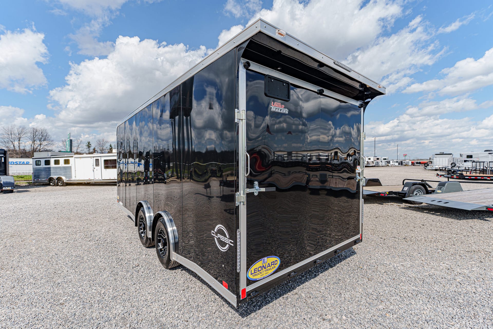 Large black enclosed dual-axle trailer parked on a gravel lot with chrome trim and a raised roof edge, reflecting the cloudy sky and surroundings in its glossy surface