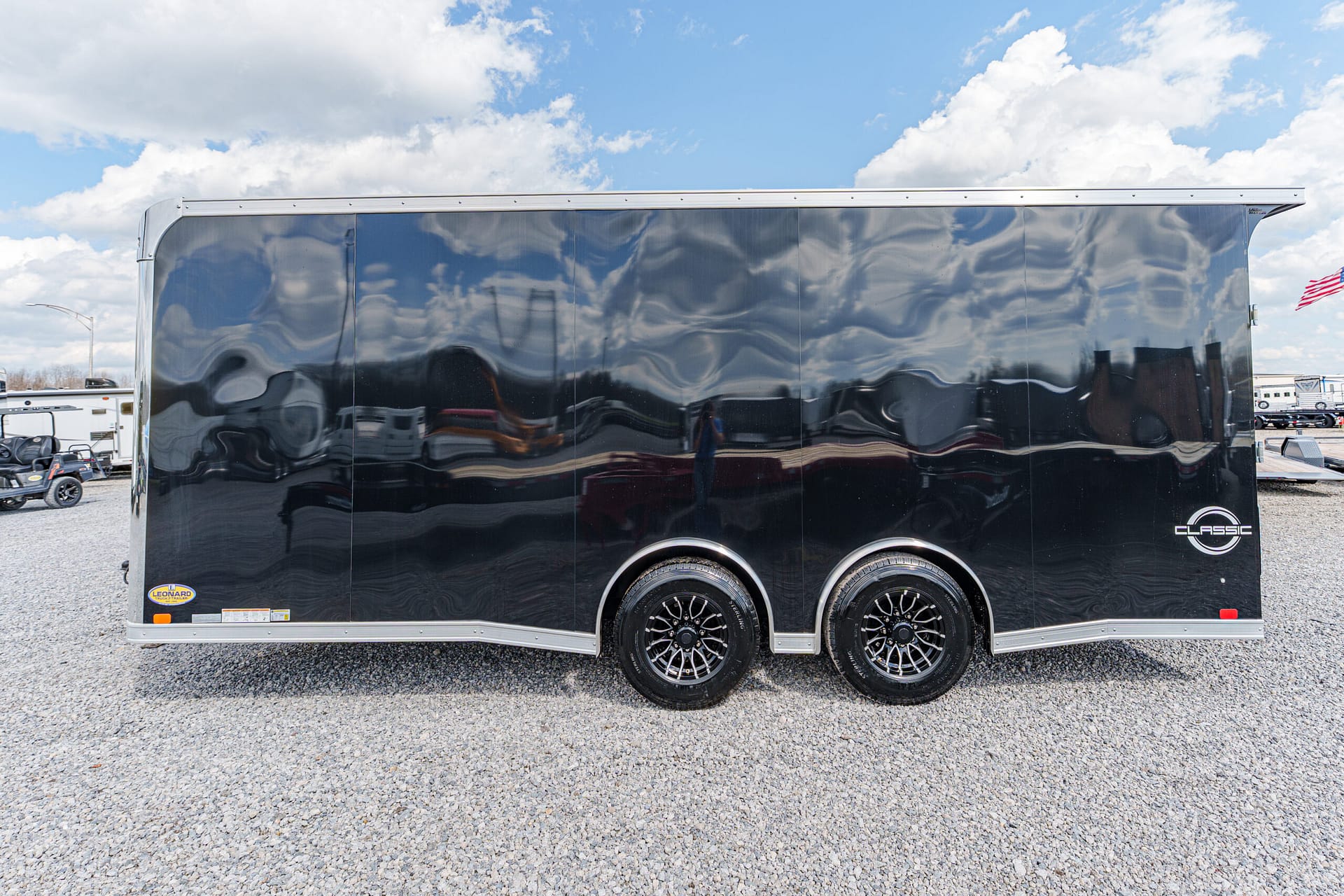 Black enclosed cargo trailer with two axles parked on a gravel lot under a blue sky with clouds.
