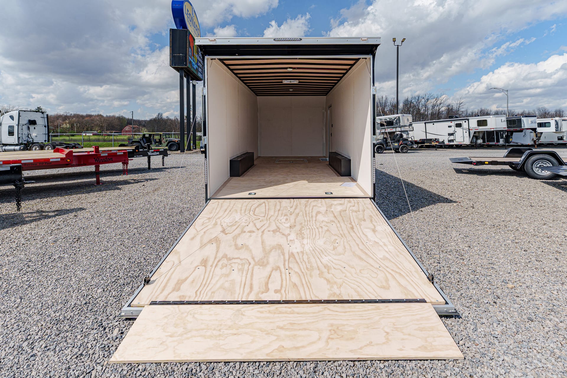 Open cargo trailer with plywood ramp lowered, empty interior and two black benches along the sides on a gravel lot.