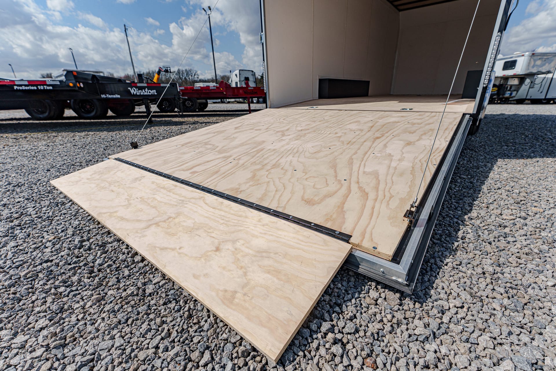 Open cargo trailer with a plywood ramp extended onto a gravel lot; interior space visible for loading.