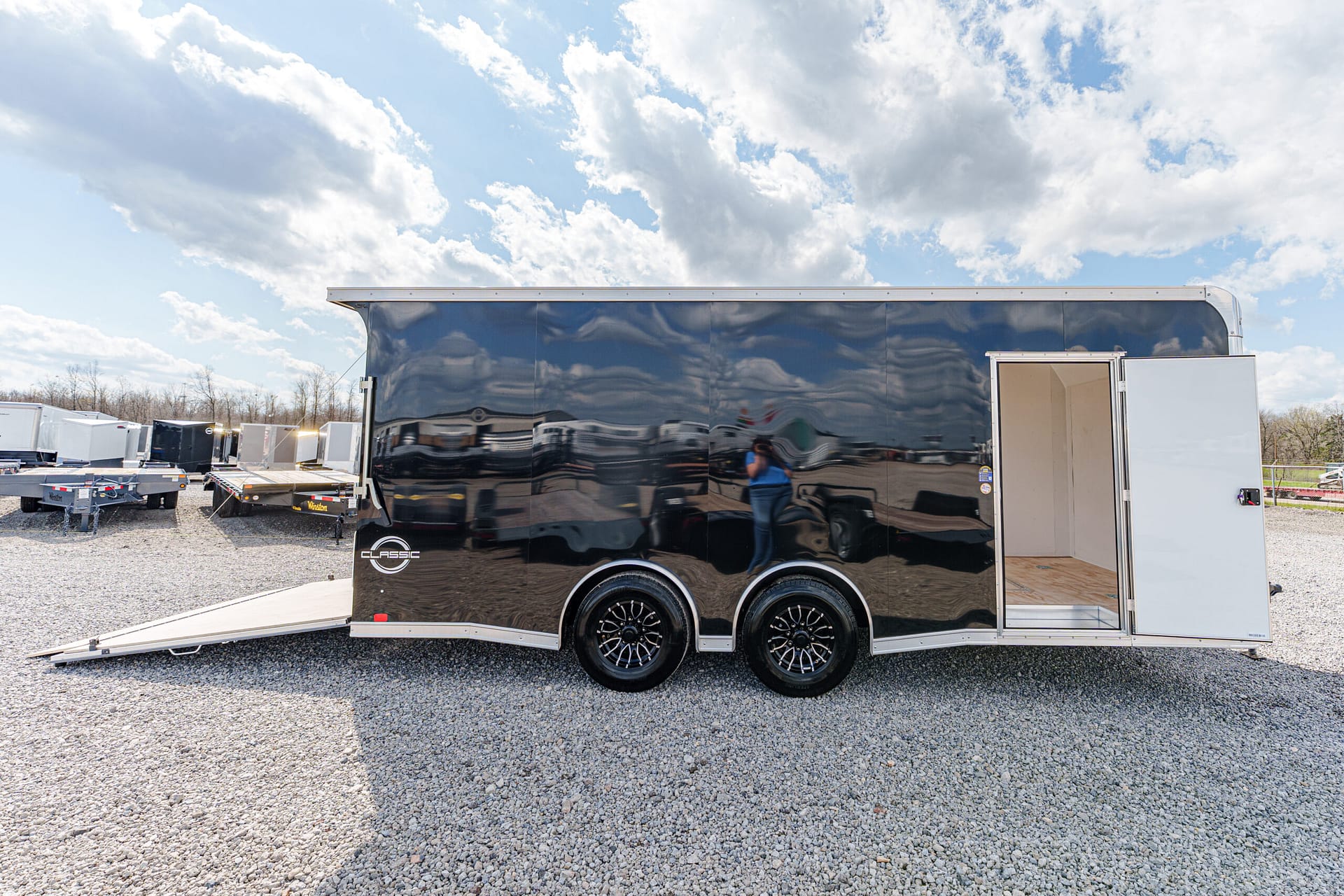 Glossy black enclosed cargo trailer with dual axles, open side door and lowered ramp on a gravel lot under a cloudy sky nearby other trailers photographed.