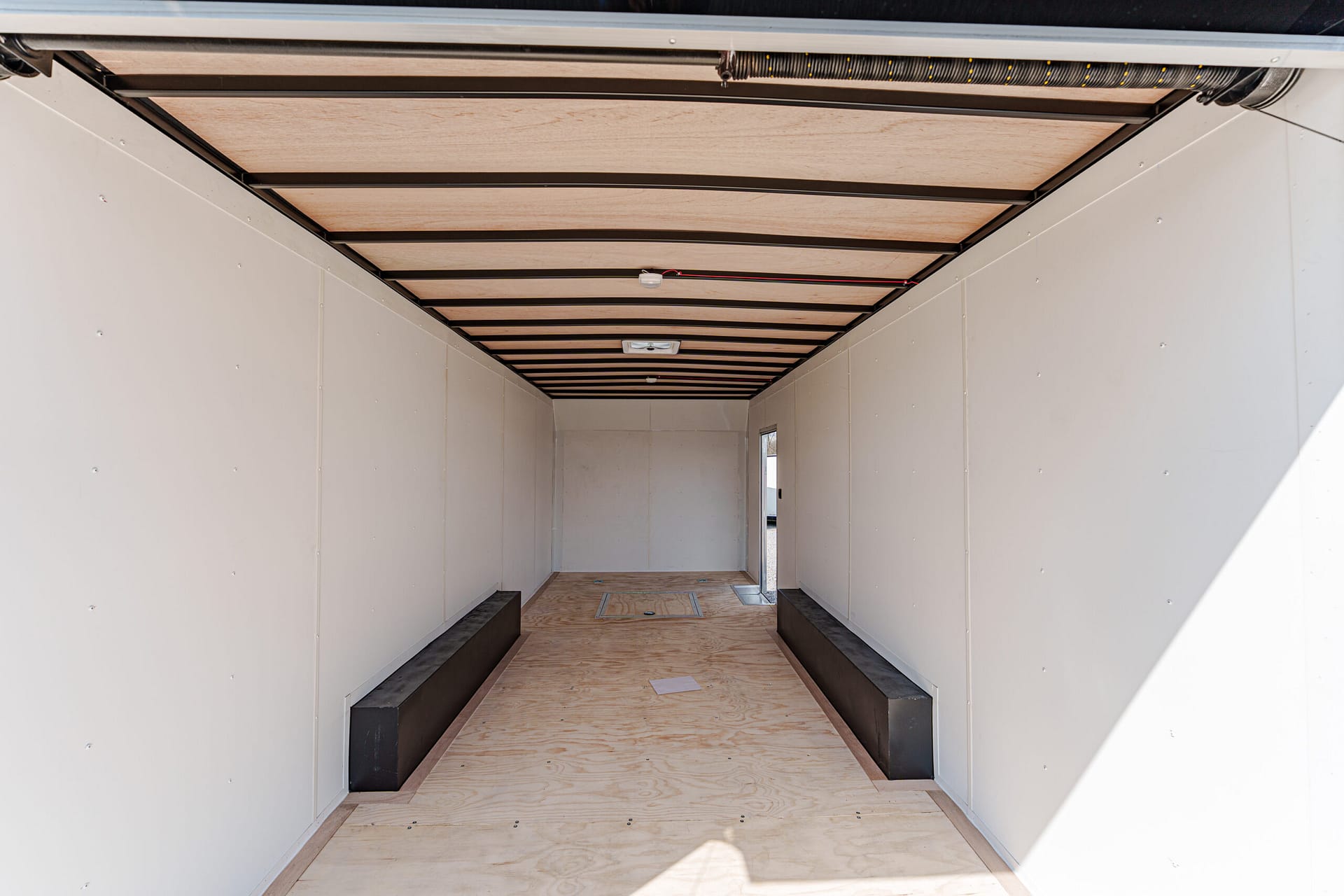 Empty trailer interior with white walls, plywood floor, and black benches along both sides.