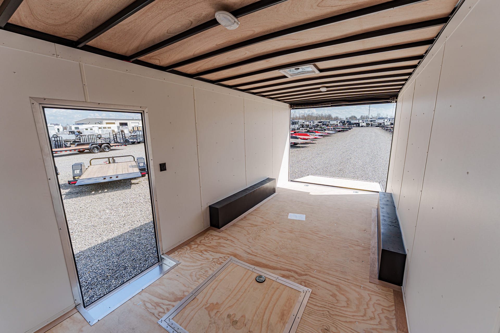 Empty trailer interior with plywood floor and black benches, open to a boat-trailer yard outside.