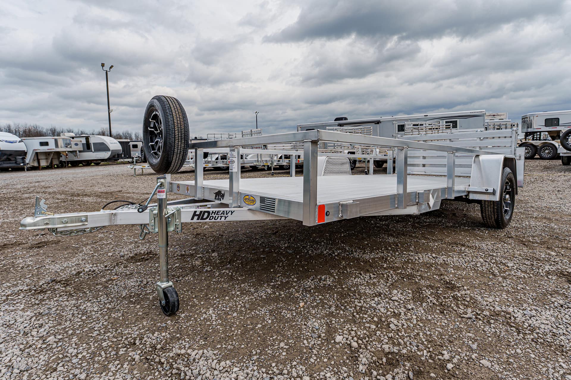 Aluminum flatbed utility trailer with a spare tire mounted at the front in a gravel lot, ready to tow.