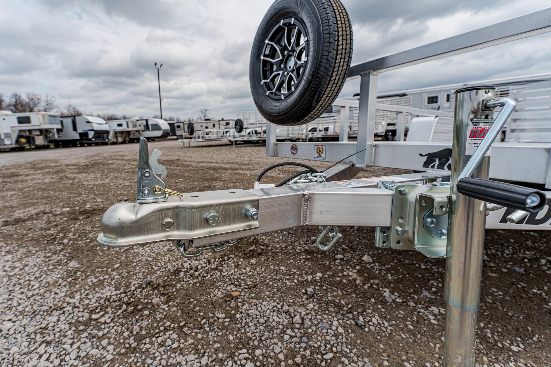 Close-up of a trailer hitch with a wheel suspended above, in a gravel RV storage yard in the background.