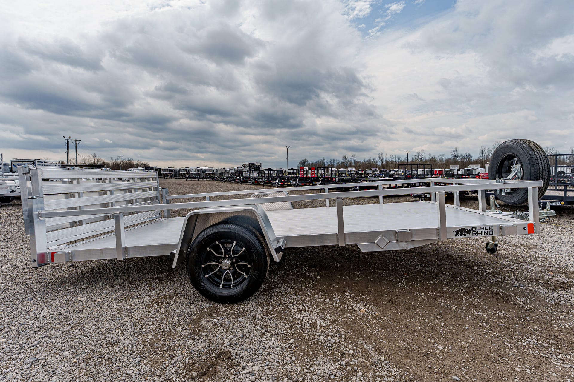 Aluminum flatbed utility trailer with wheel and spare tire, parked in a gravel lot among other trailers under a cloudy sky