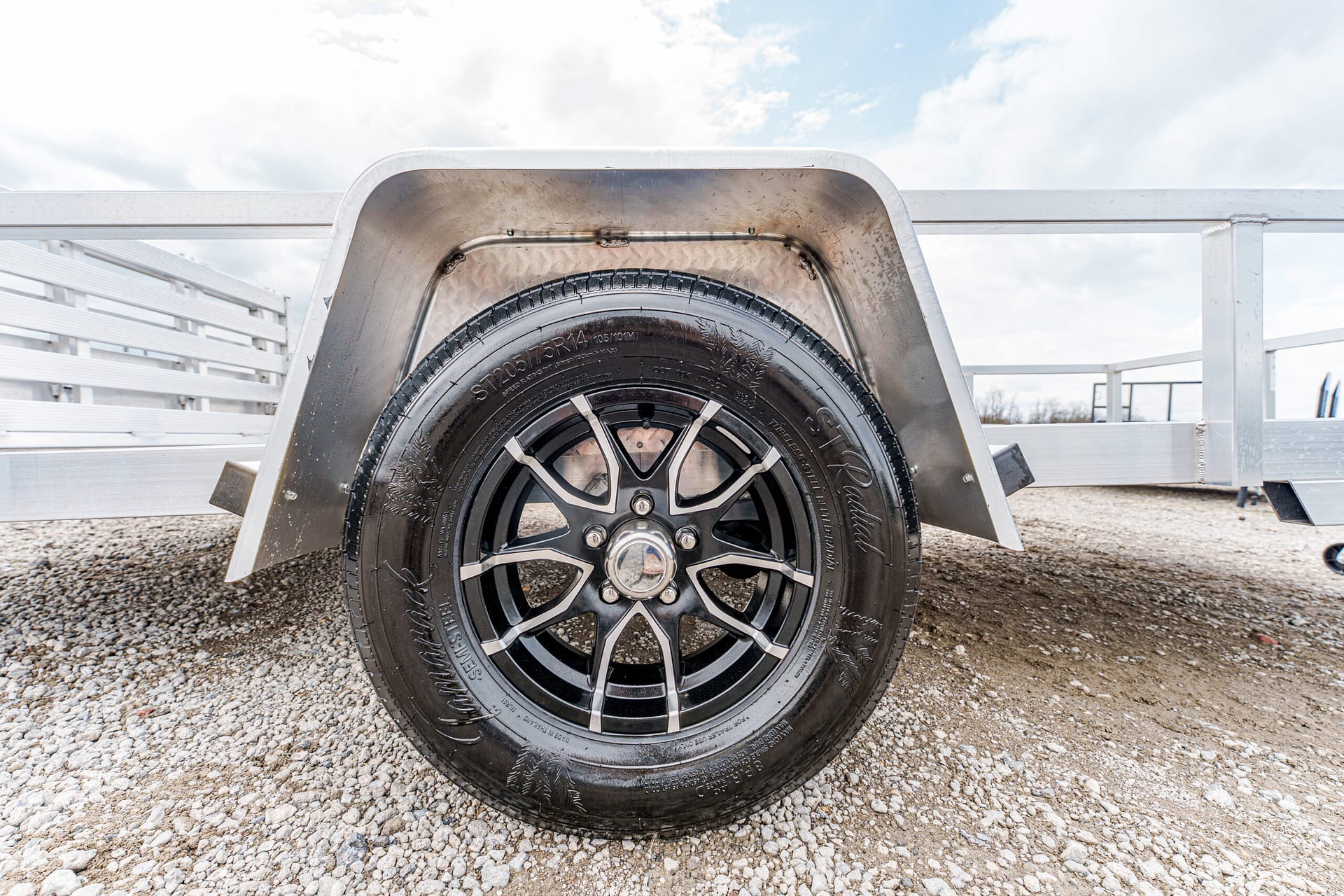 Wheel of a utility trailer with black alloy rim and tire, inside a metal fender on a gravel lot