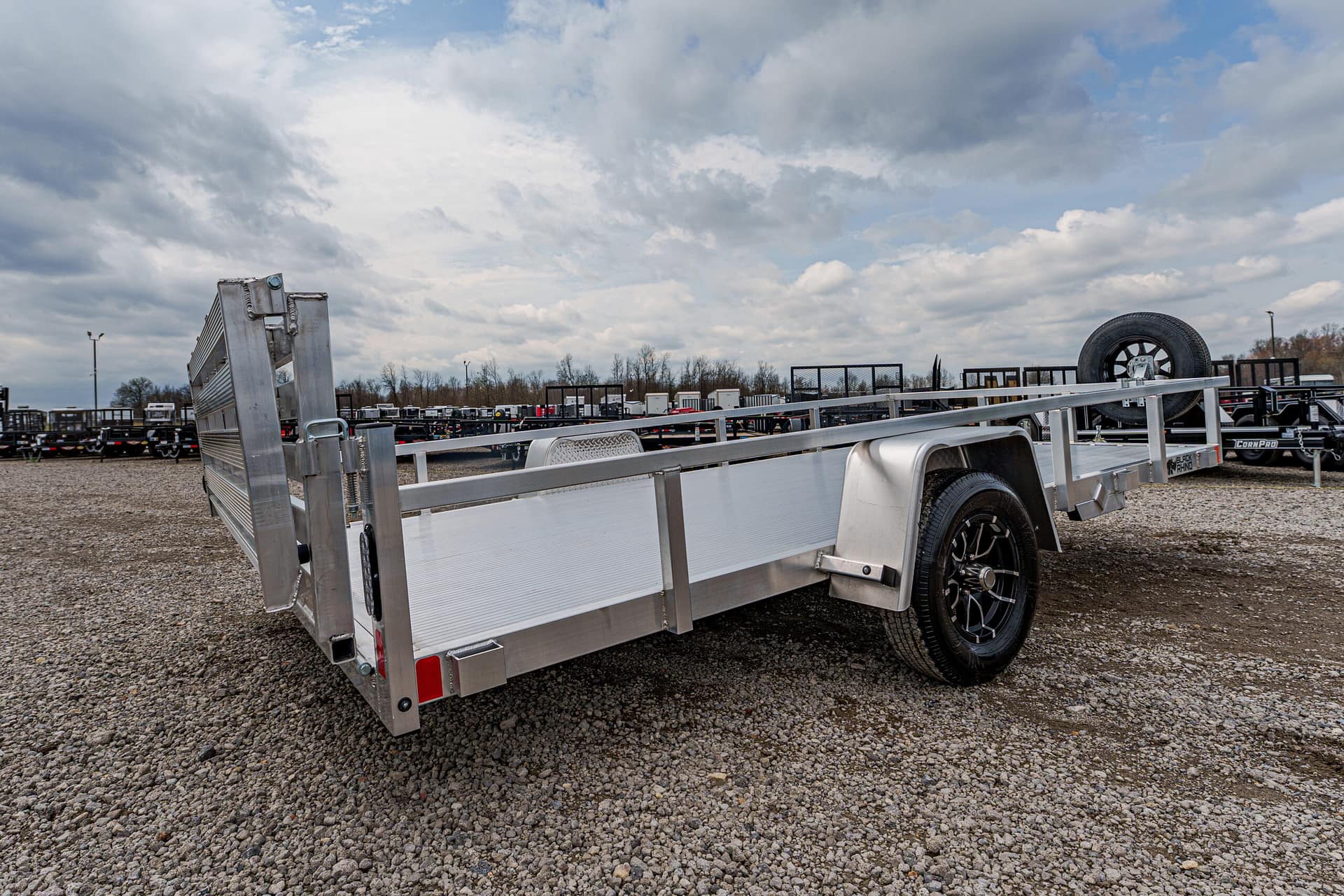 Aluminum utility trailer with side rails and ramp gate parked on a gravel lot, spare tire visible in background.