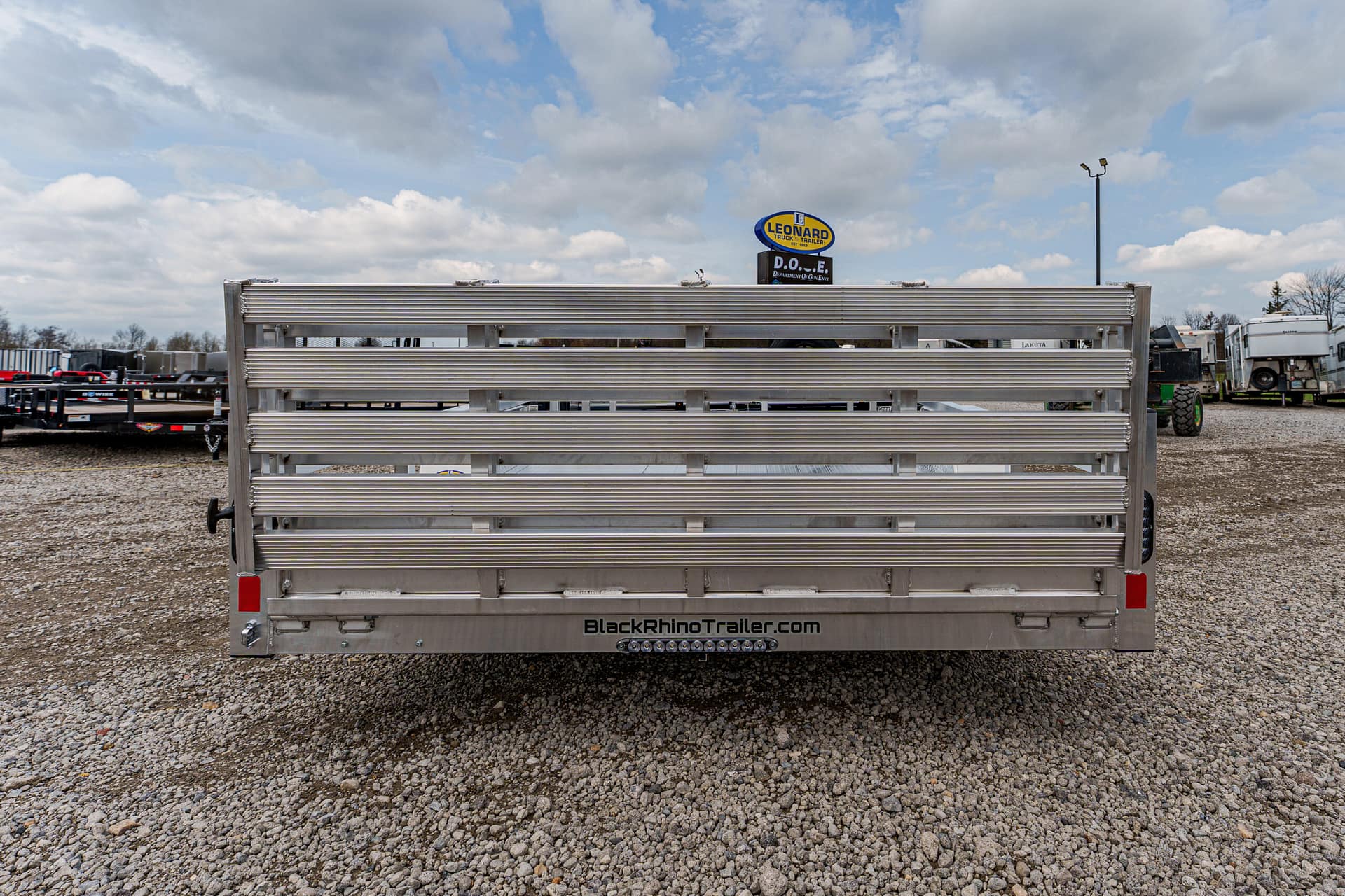 Rear view of a silver aluminum trailer with horizontal slats and BlackRhinoTrailer.com on the bumper, parked on a gravel lot under a cloudy sky.