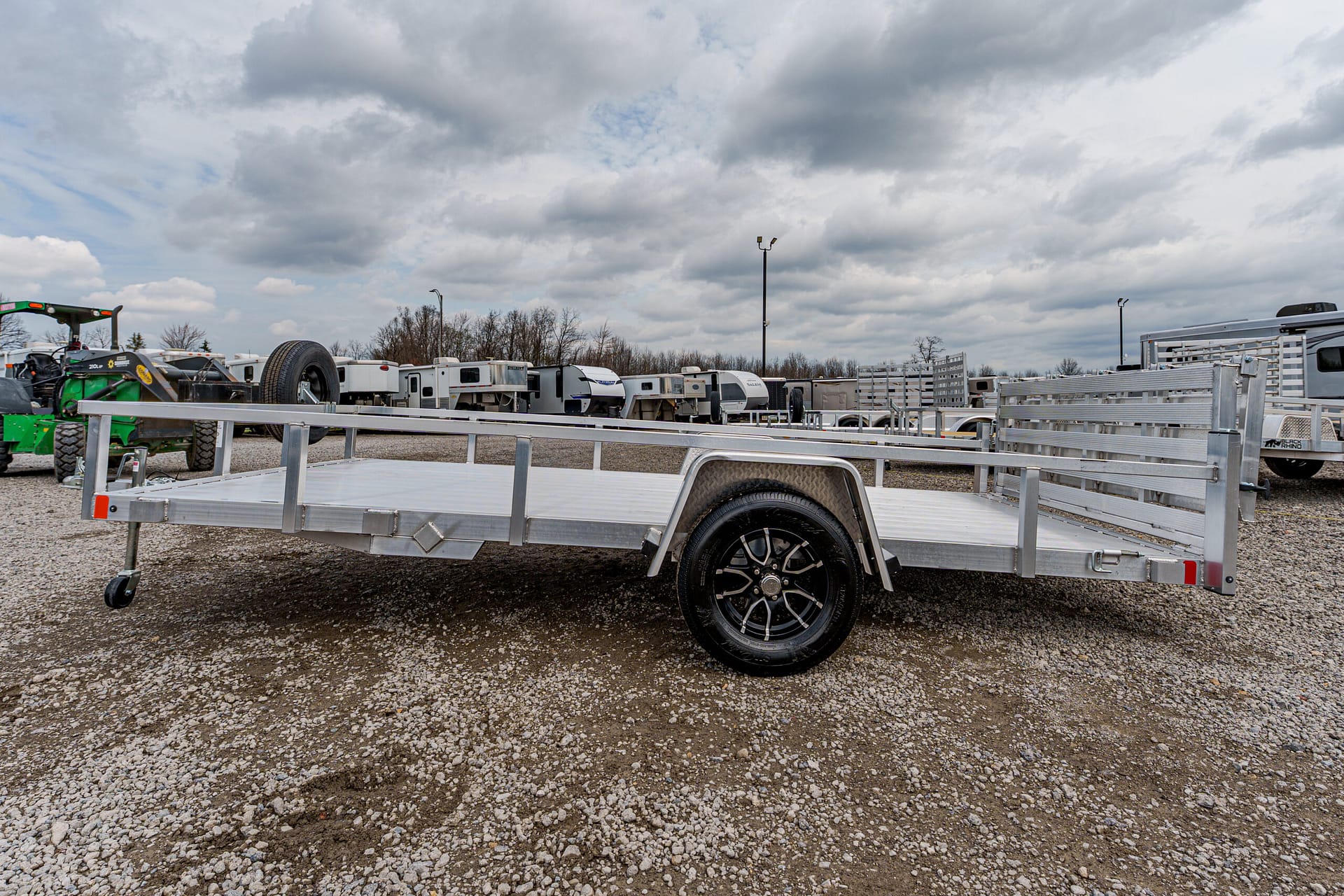 Aluminum flatbed trailer with a single axle and spare tire, parked on a gravel lot among other trailers in the background.