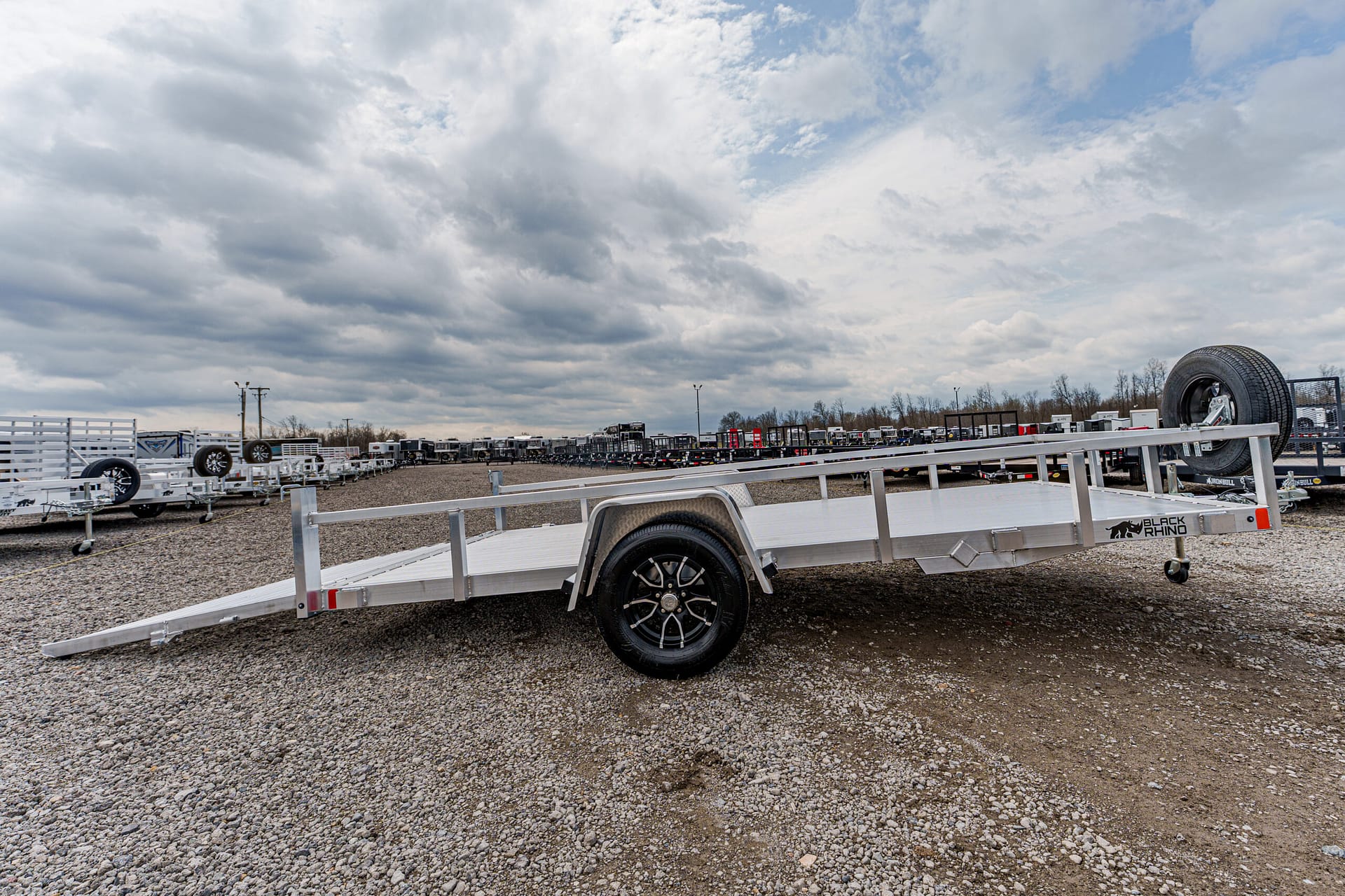 Aluminum flatbed trailer with ramp extended, parked in a gravel lot among many trailers.