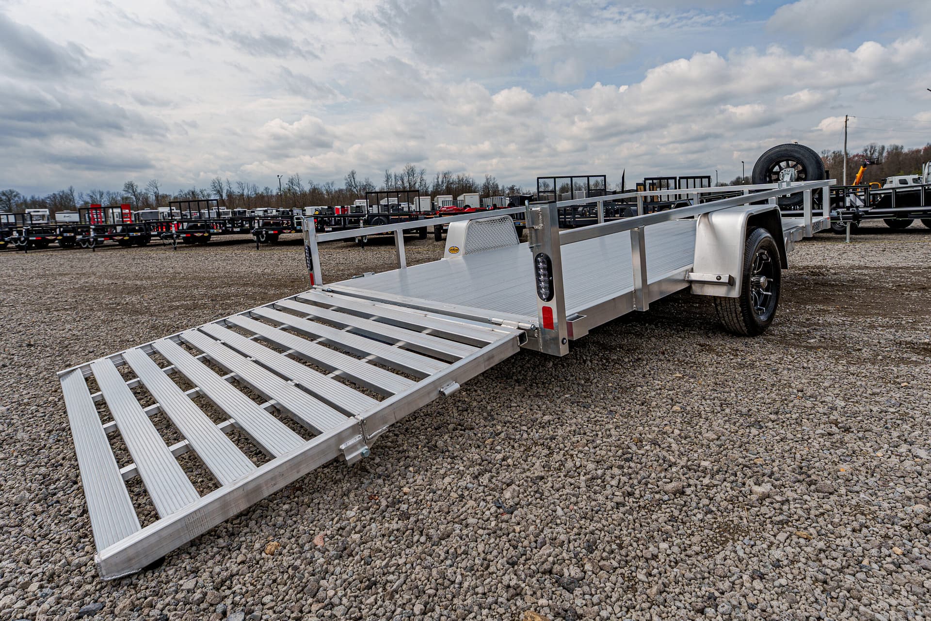 Aluminum flatbed trailer with extended loading ramp in a gravel lot; other trailers visible in the background.