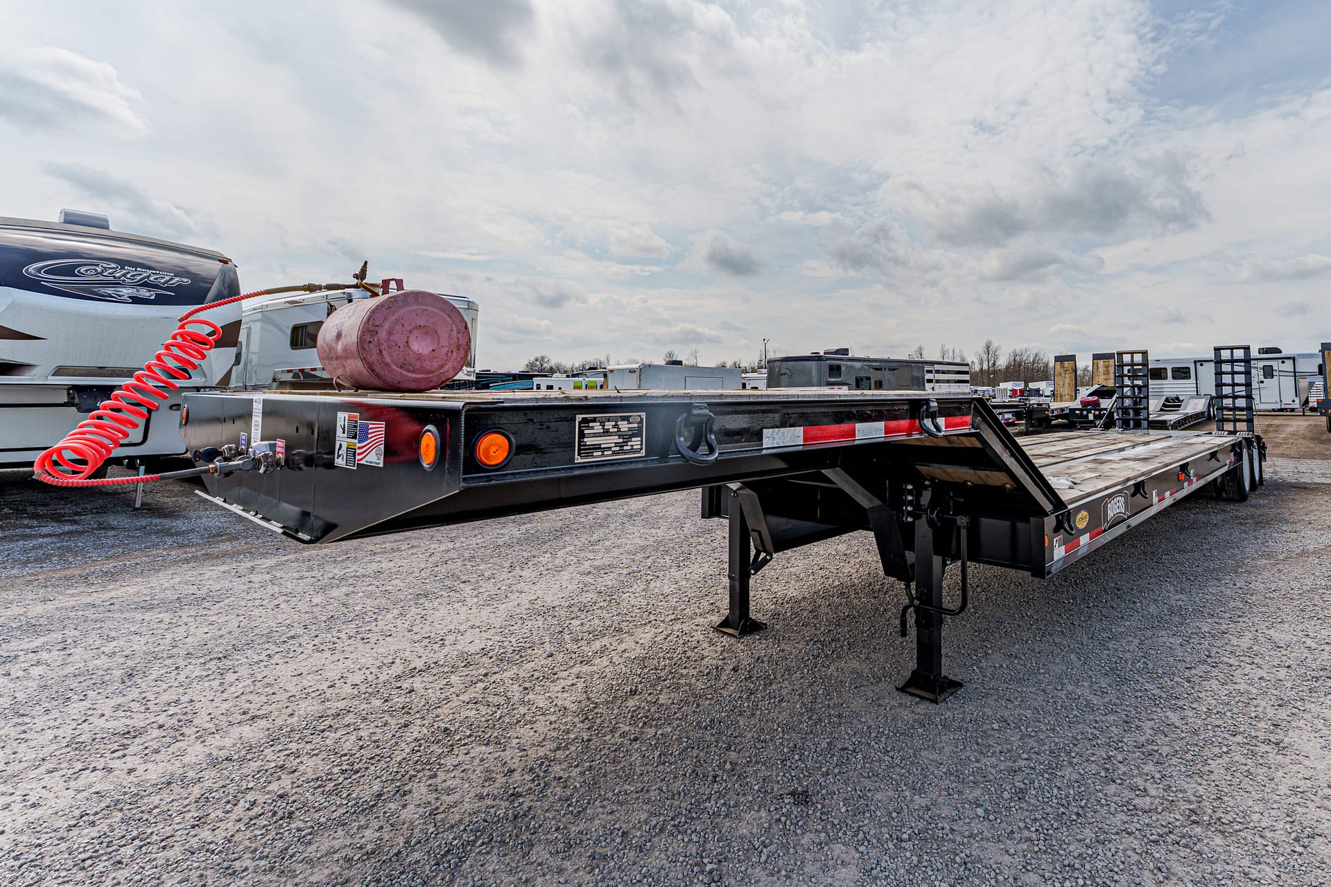 Gooseneck flatbed trailer with a red propane tank and coiled orange hose on top, parked on a gravel lot among RVs and trailers.