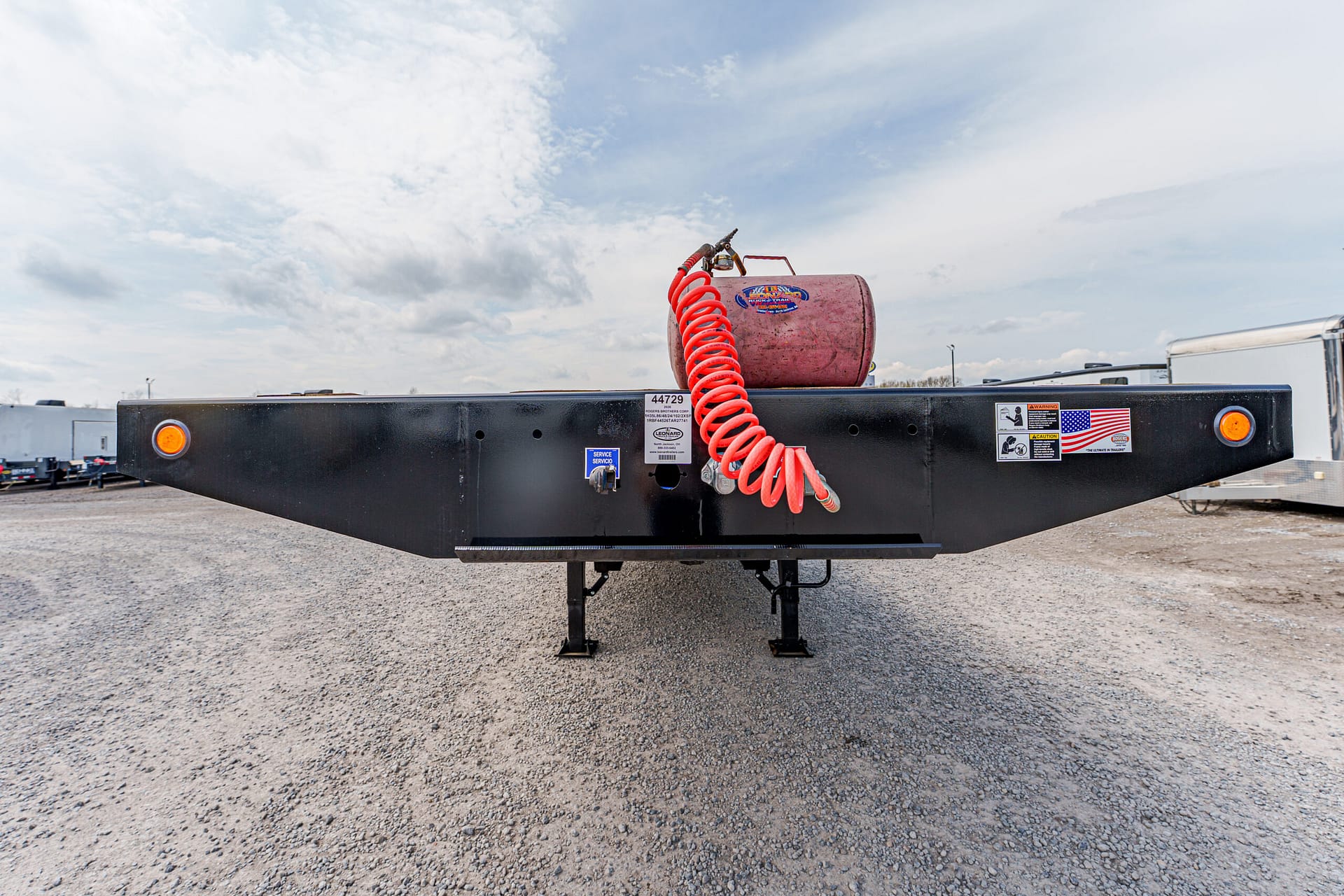 Black flatbed trailer with a red coiled hose and a pink gas canister mounted on top, parked on a gravel lot under a cloudy sky.