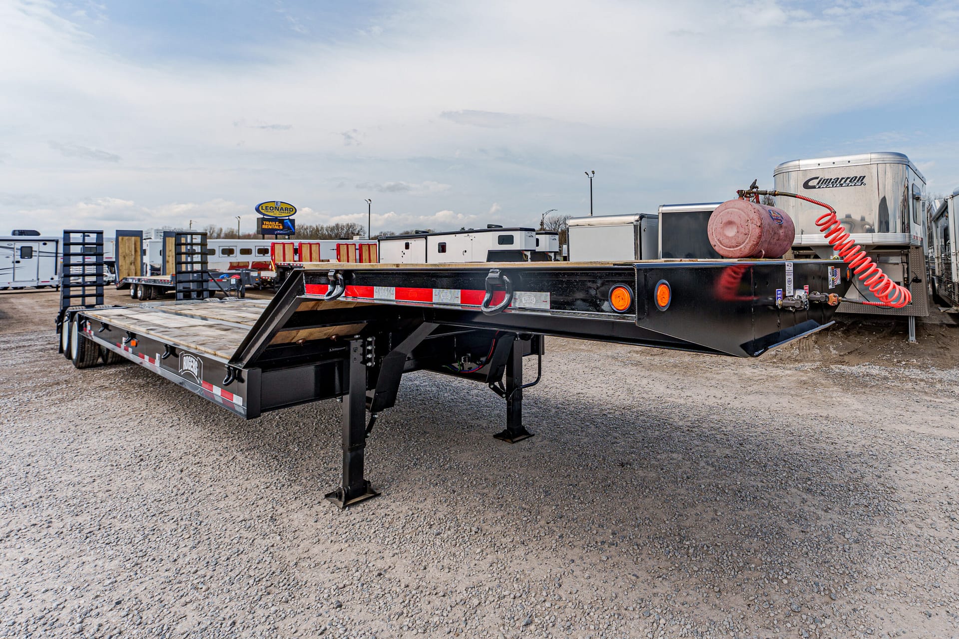 Black flatbed trailer on a gravel lot, with ramps down and orange reflectors, surrounded by other trailers and trucks in the yard.