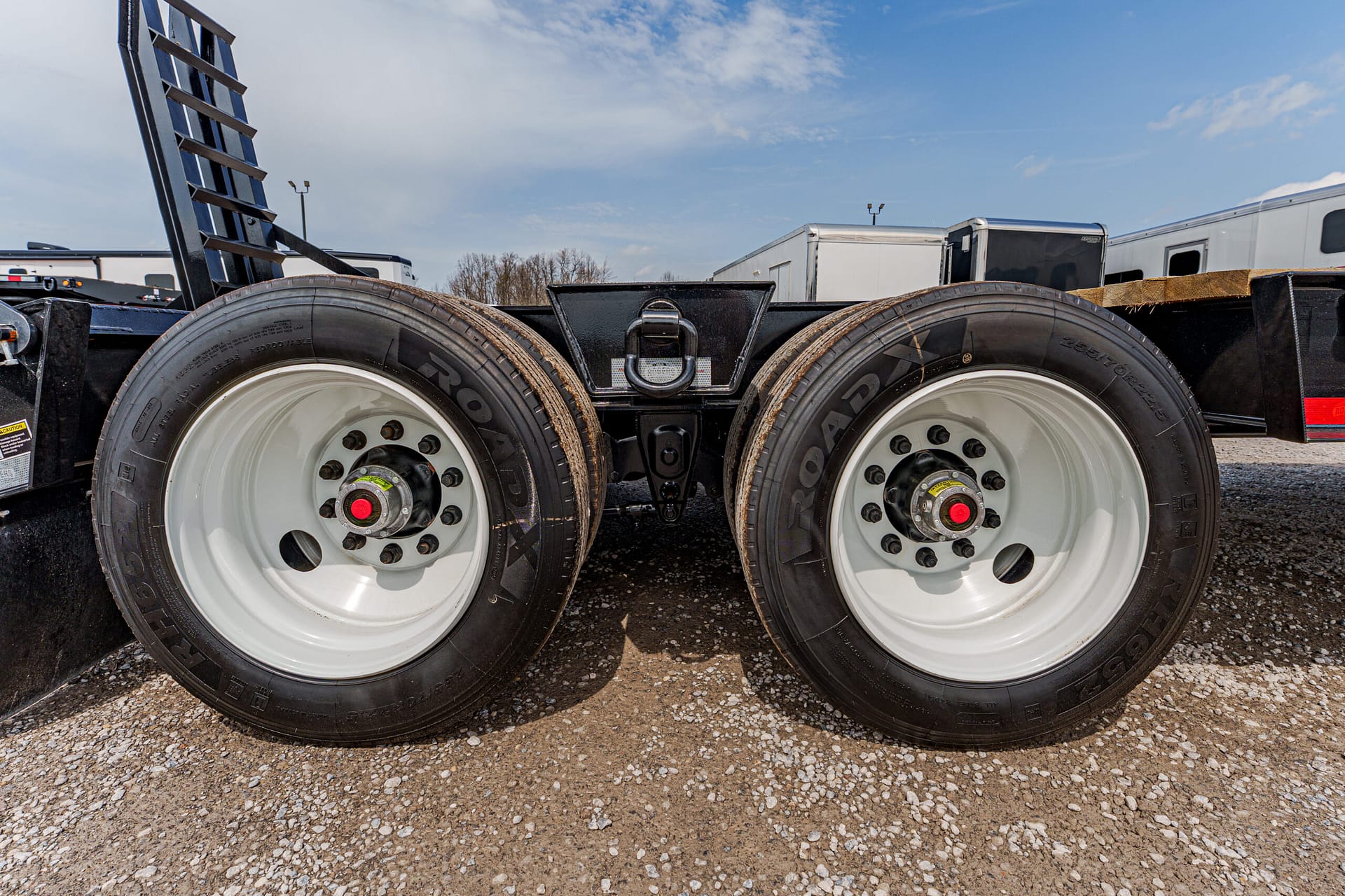 Dual heavy-duty trailer wheels with white rims on a gravel lot, showing red center caps and black undercarriage under a portable trailer setup.