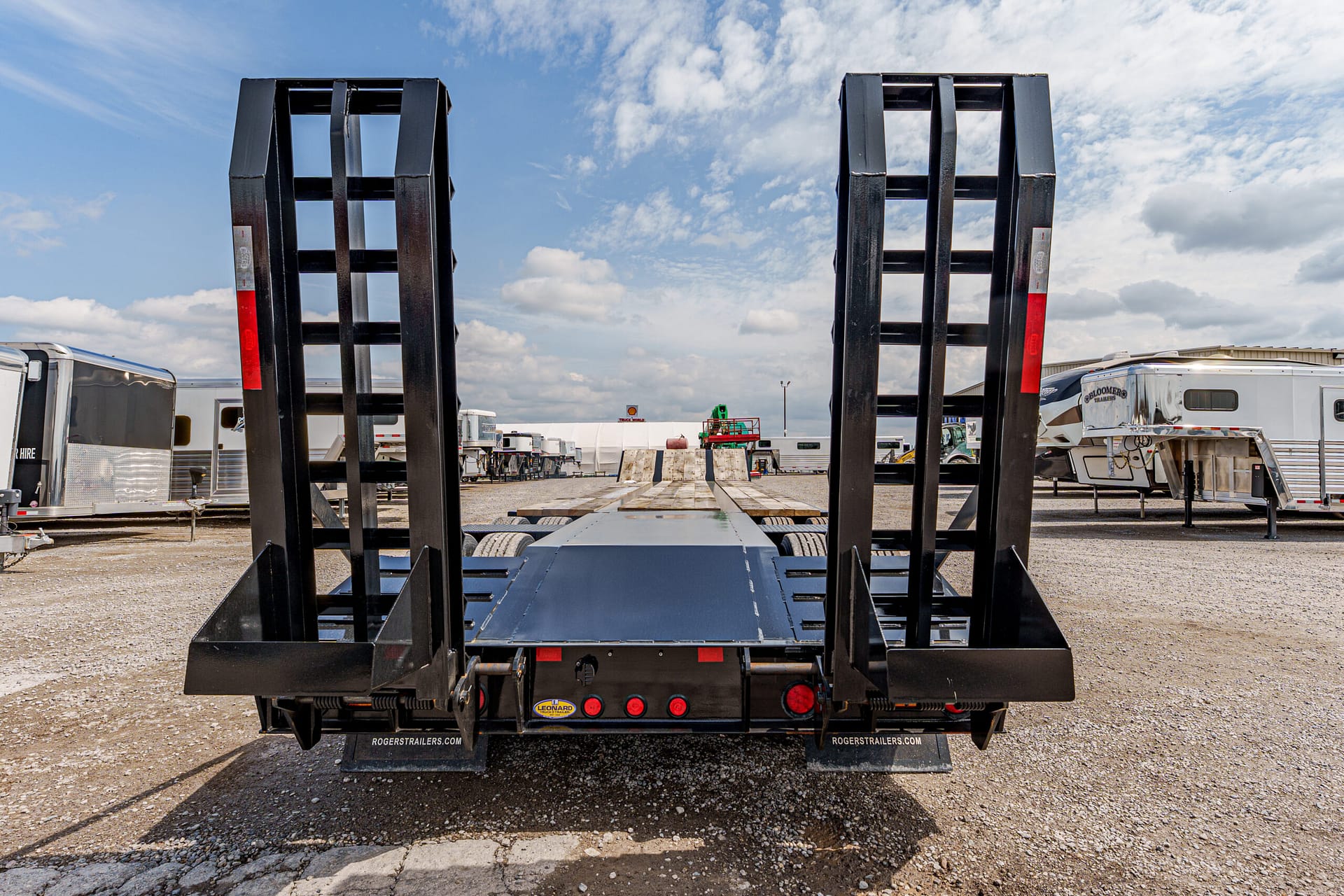 Rear view of a black flatbed trailer with two tall loading ramps raised, in a gravel trailer yard with other trailers nearby, under a blue sky.