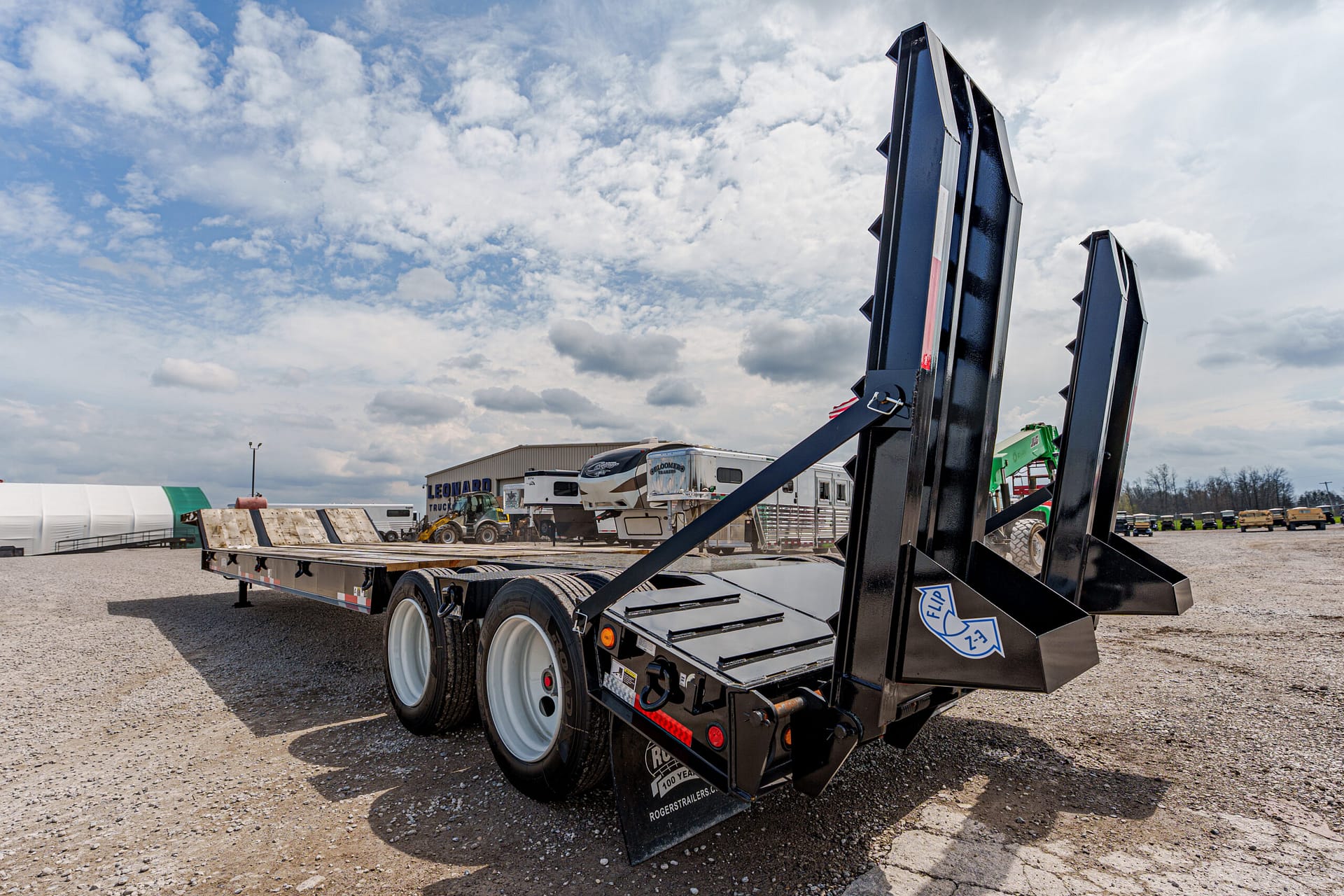 Black dual-ramps trailer with raised loading ramps on a gravel lot, with construction equipment and RVs in the background.