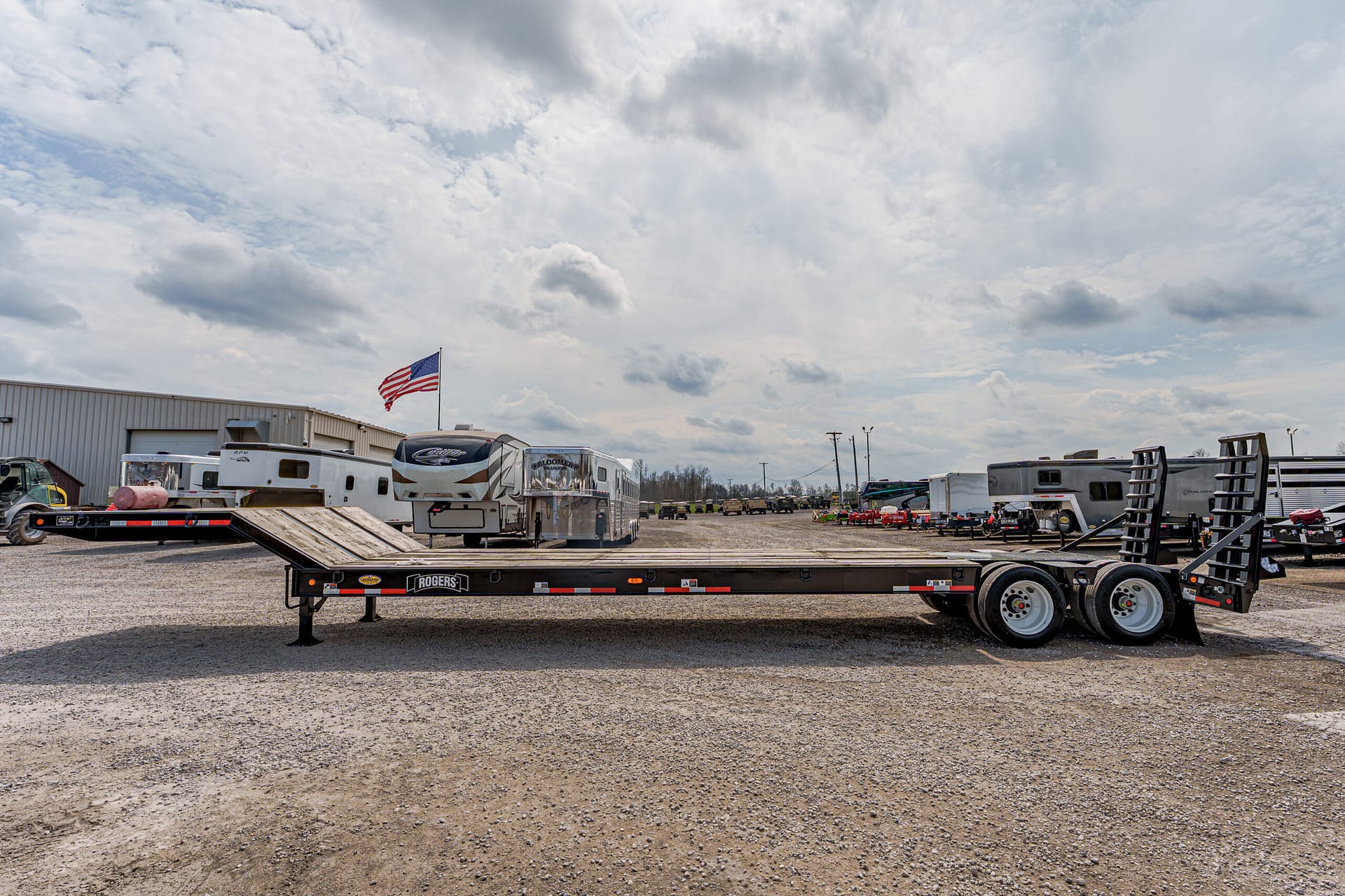 Long flatbed trailer in a gravel lot with RVs and camping trailers, an American flag flying nearby under a cloudy sky.