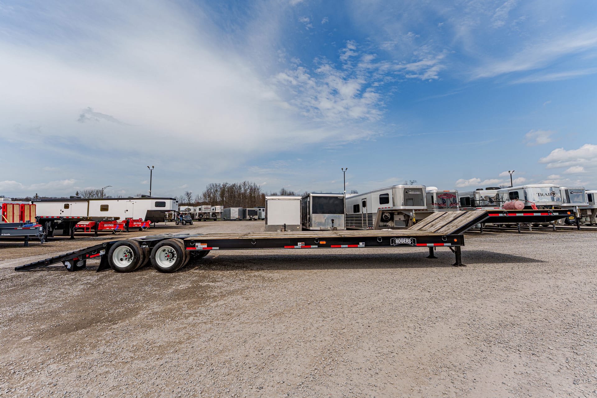 Black flatbed trailer with ramps lowered in a gravel lot, other trailers parked behind it on a sunny day