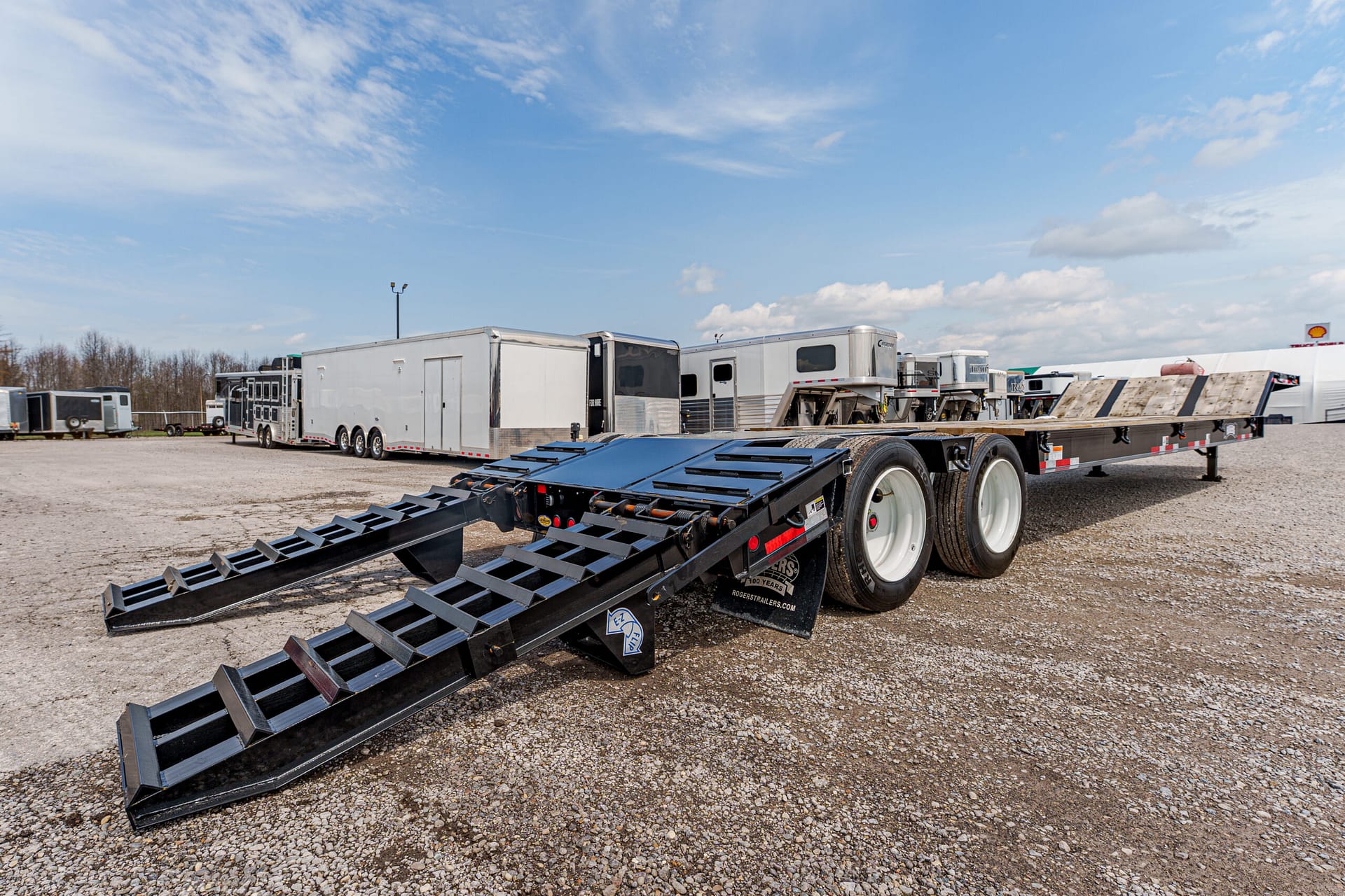 Flatbed trailer with heavy steel loading ramps extended on a gravel lot, with other trailers in the background.