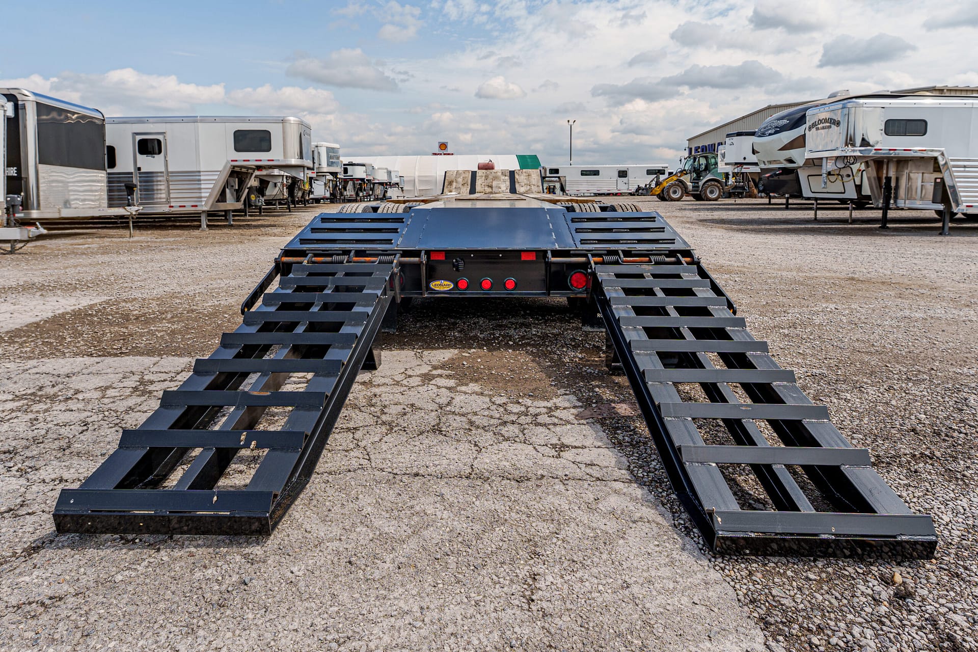 A wide truck trailer with two black ramps extended on a gravel lot, surrounded by other trailers and equipment under a cloudy sky.