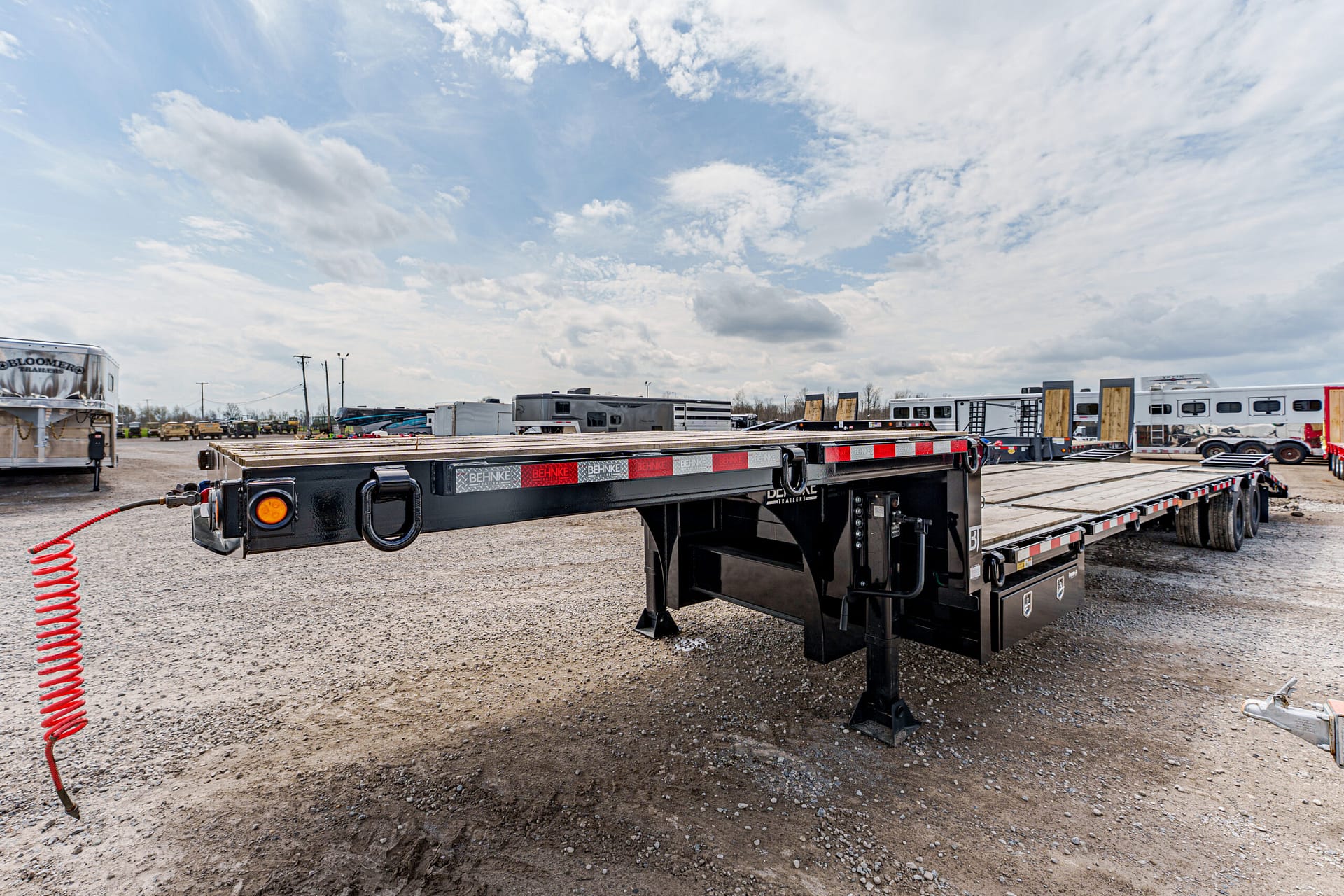 Low-profile black flatbed trailer in a gravel lot with other trailers and trucks in the background under a cloudy sky