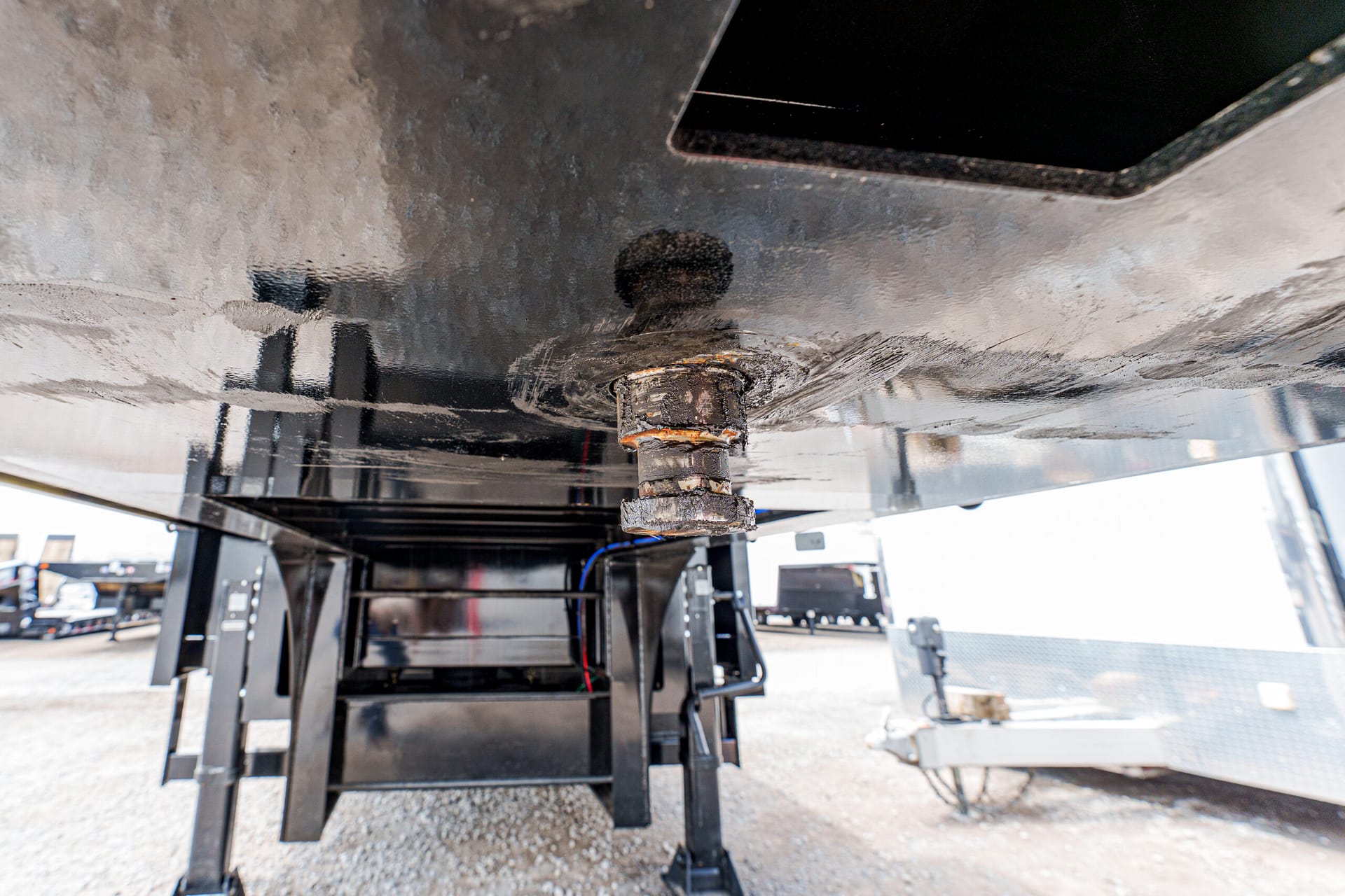 Rusty trailer hitch connection seen from underneath, with jack stands supporting the trailer and other trailers in the background.