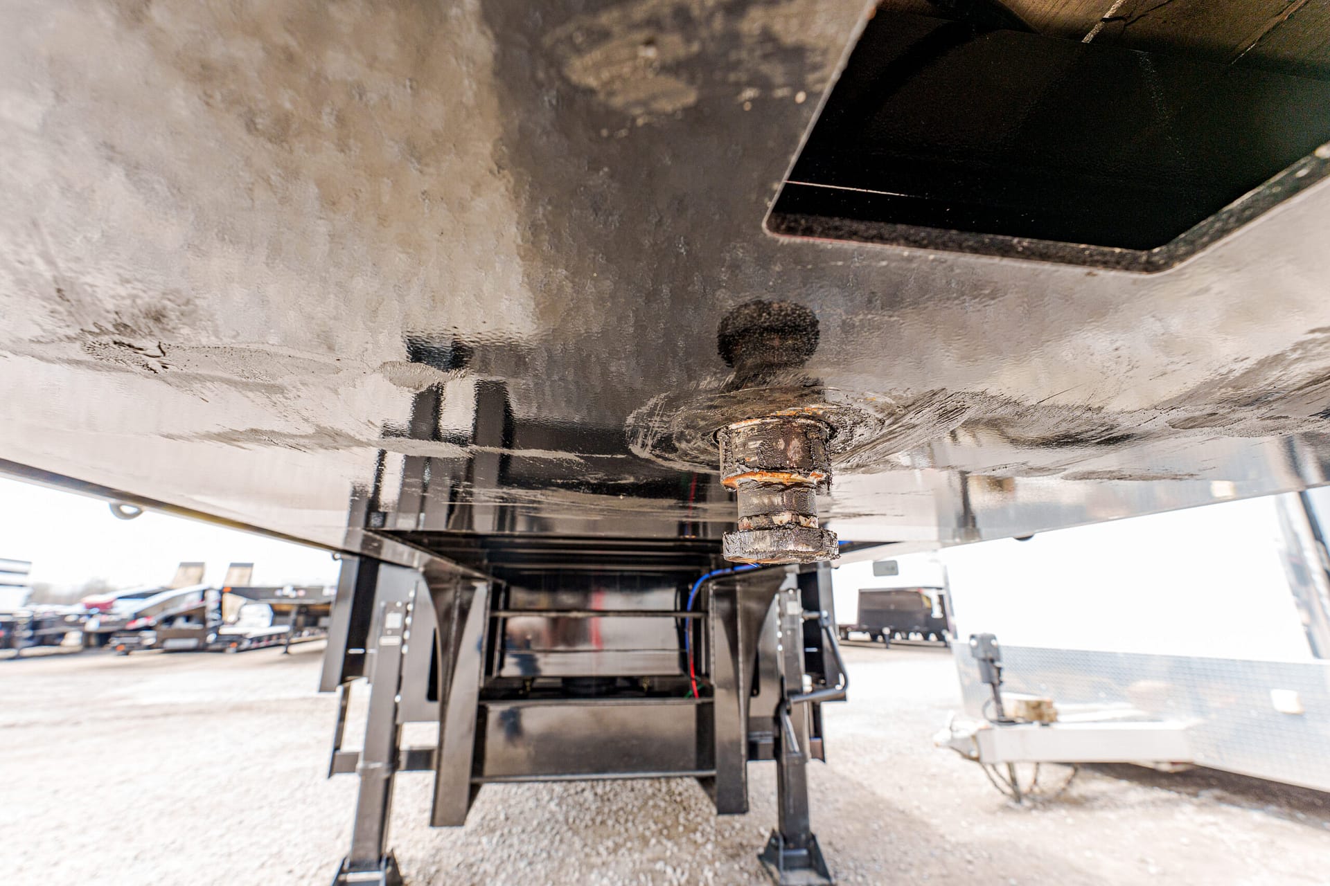 underside of a trailer with its stabilizing legs down, showing a rusty coupling pin and dirty undercarriage in a gravel lot.