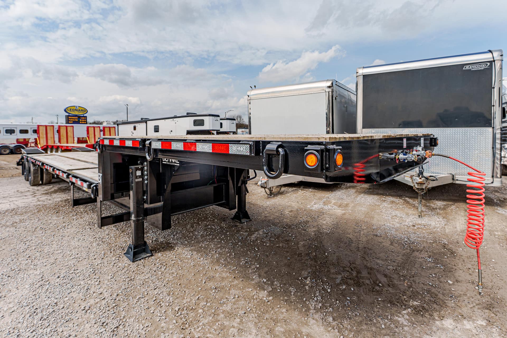 Black flatbed trailer on a gravel lot, with orange reflectors and a red coiled air hose, in a trailer park setting.